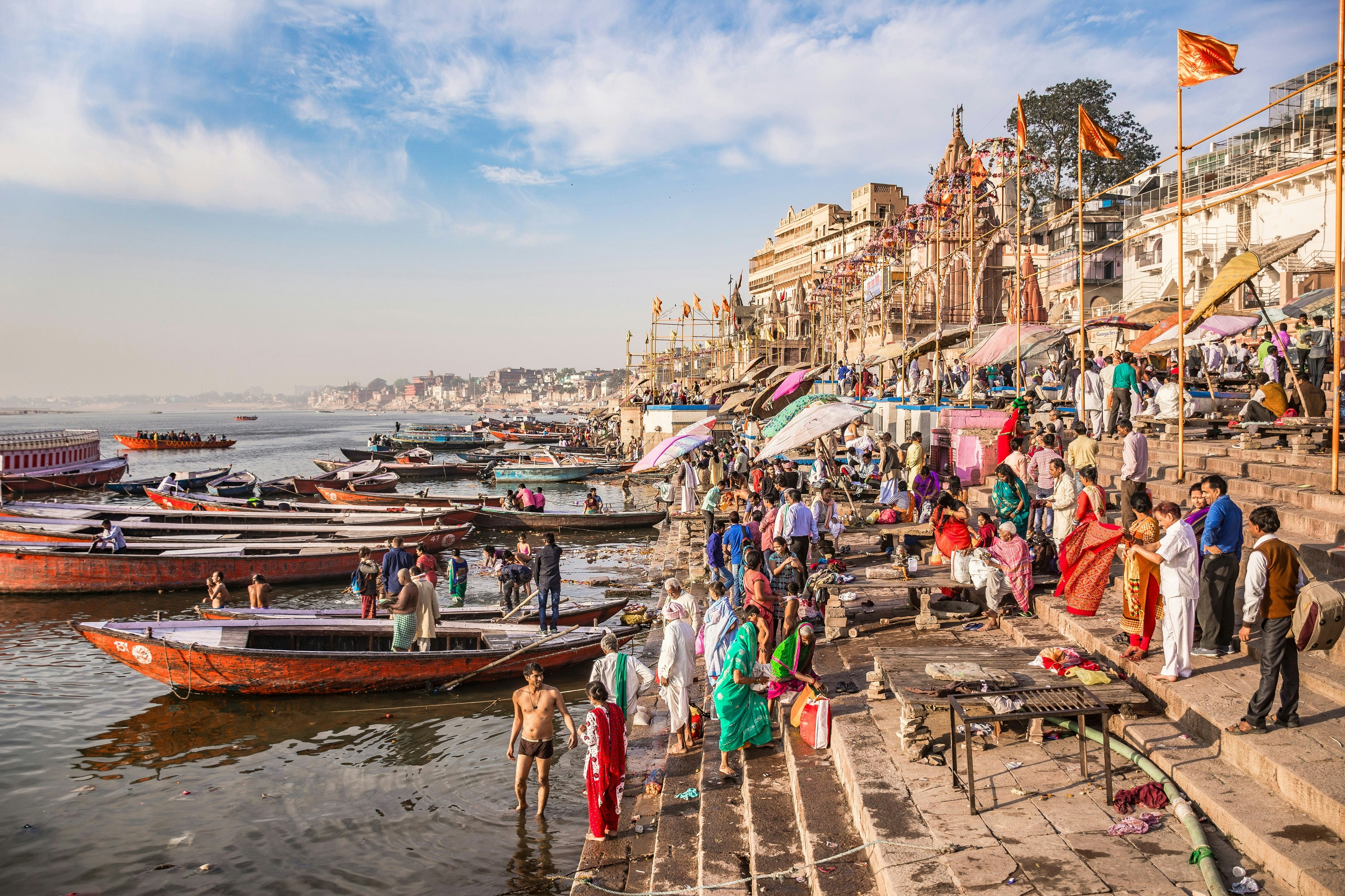 Scores of people are gathered at steps leading down to the Ganges River at Varanasi; some are standing in the water, and some are sitting in long, narrow boats moored at the water's edge.