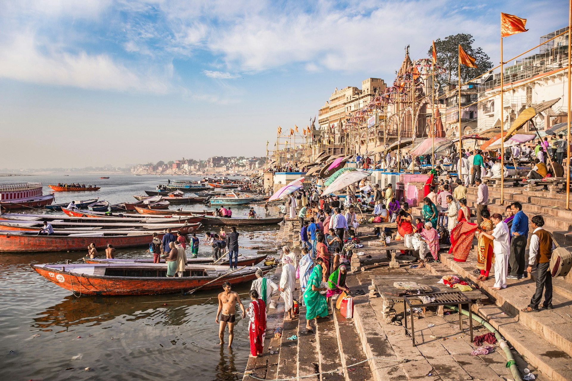Scores of people are gathered at steps leading down to the Ganges River at Varanasi; some are standing in the water, and some are sitting in long, narrow boats moored at the water's edge.