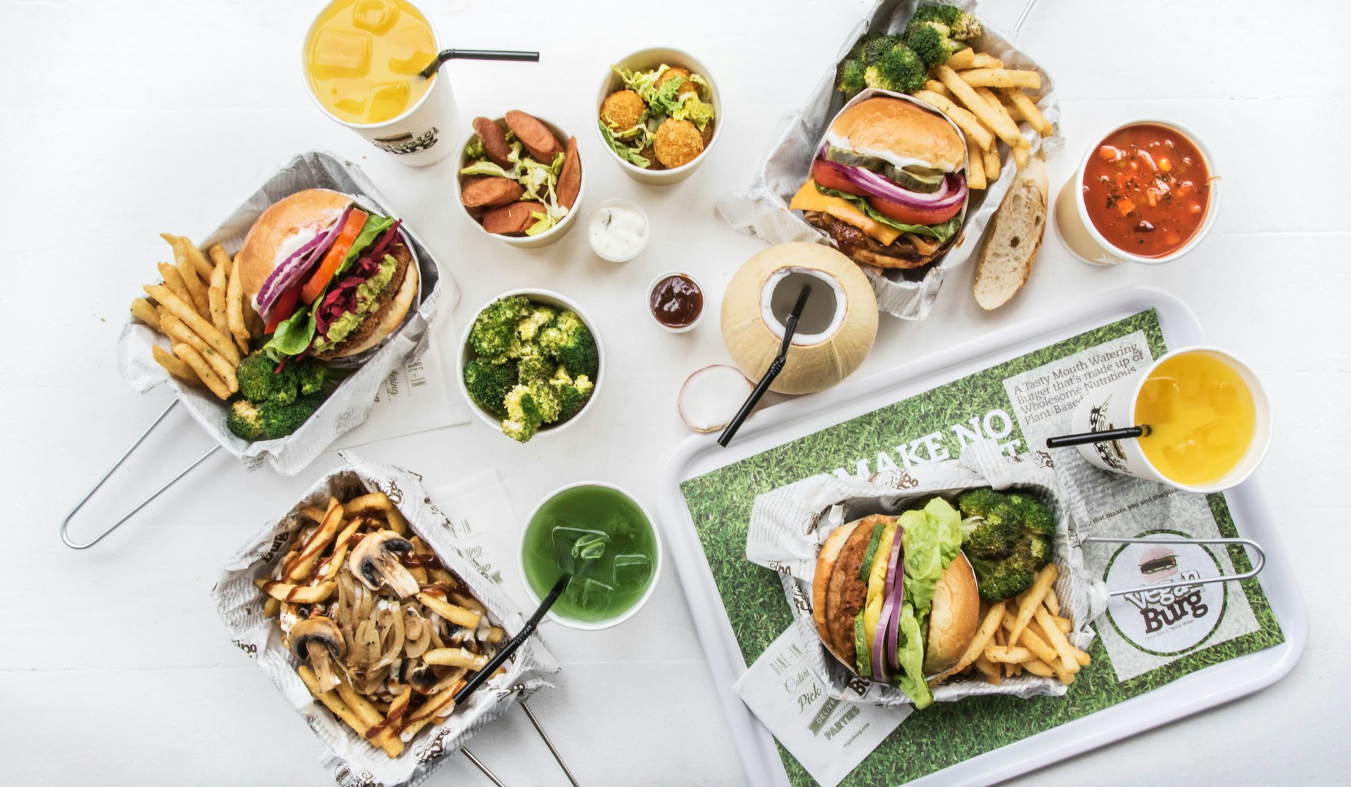 An overhead view of a table filled with dishes of vegan burgers with broccoli and french fries, small salads and a variety of cups; San Francisco Bay vegan restaurant 