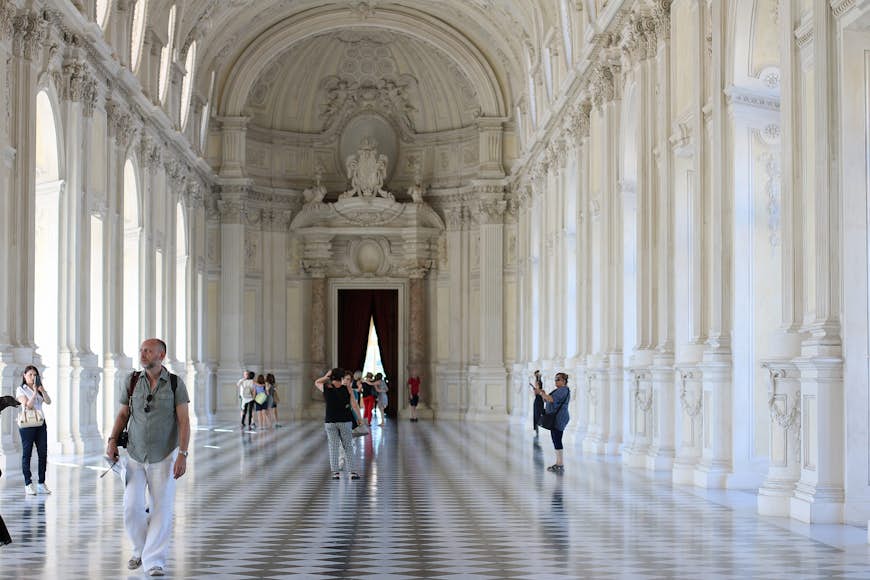 Människor som går genom Venaria Reale-palatsets vidsträckta barockhall med svartvita marmorplattor, utsmyckade vita pelare och ett välvt tak.