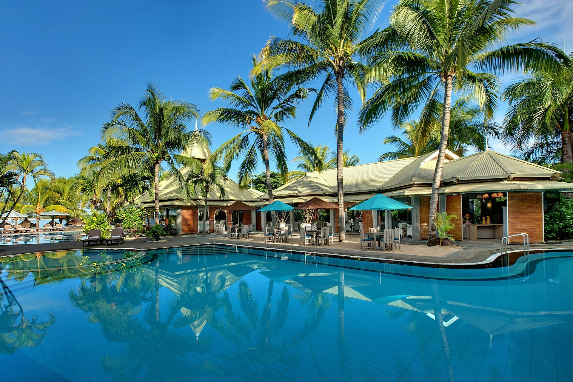 A swimming pool at the Veranda Grand Baie Mauritius, on a sunny day. Palm trees and awnings are visible poolside, along with chairs and parasols