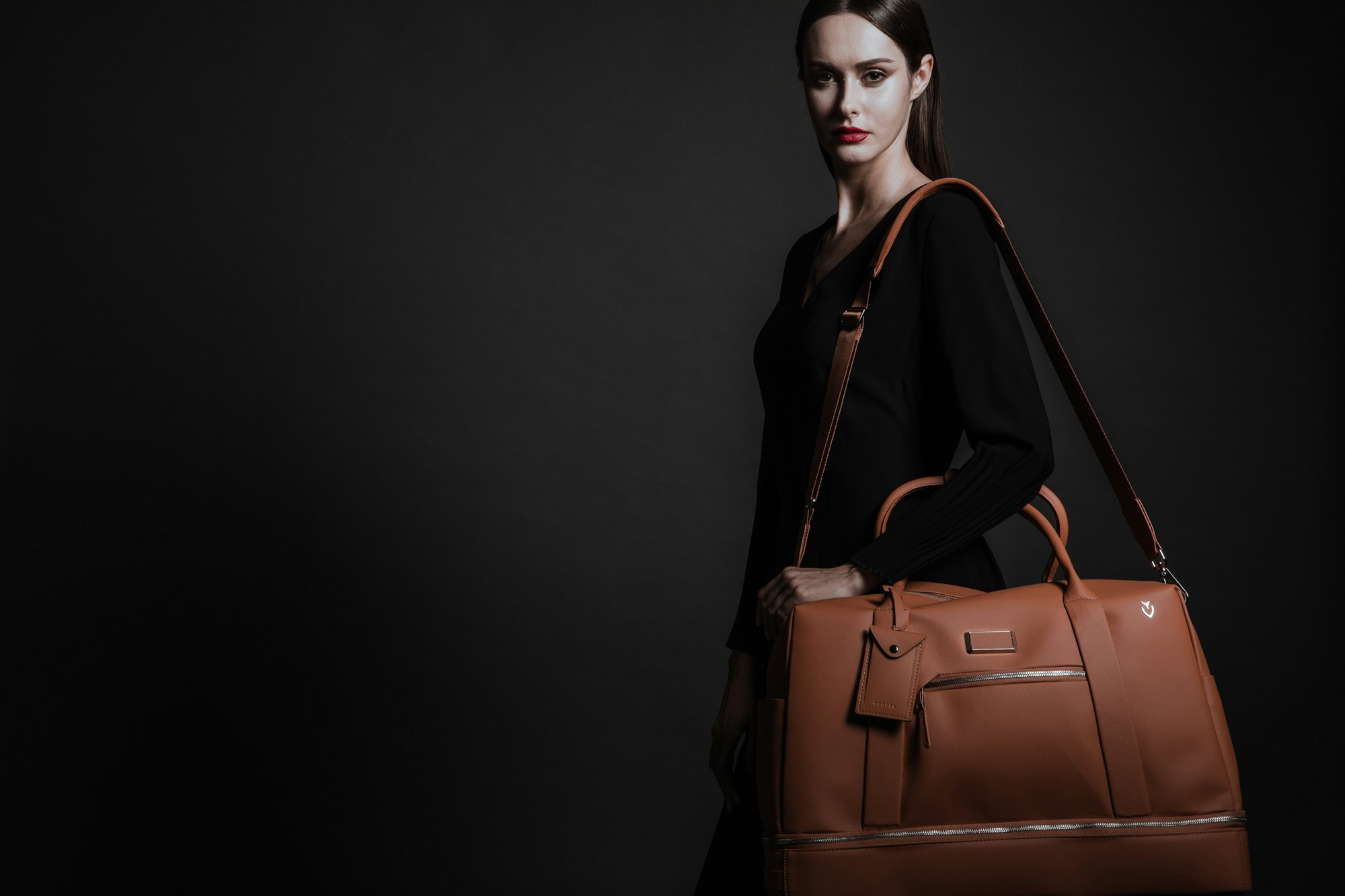 A woman in black with long hair and red lipstick, carrying the brown Vessel Signature 2.0 Boston duffel over her shoulder