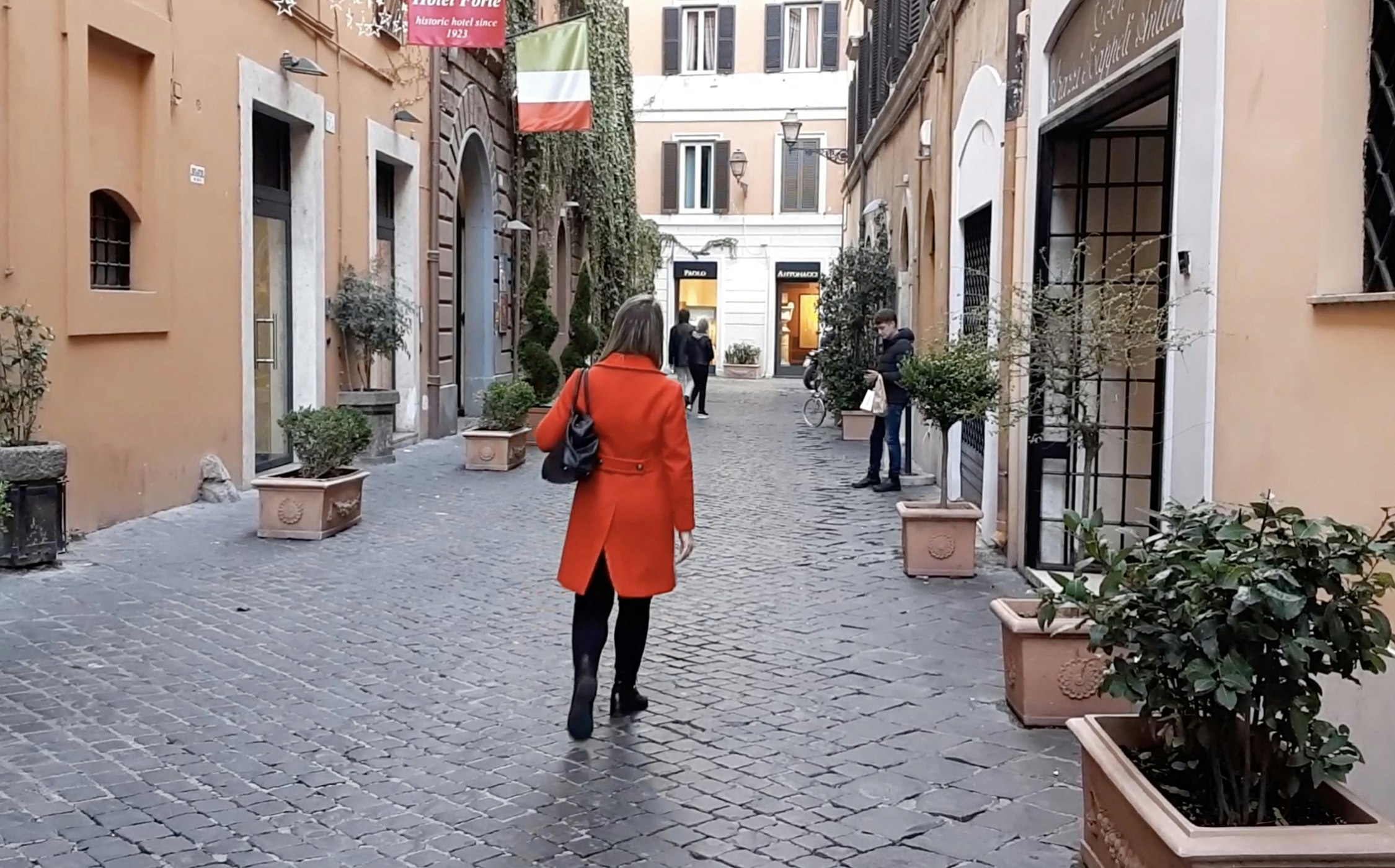 A woman in a red jacket walks down a cobbled street in Rome. 