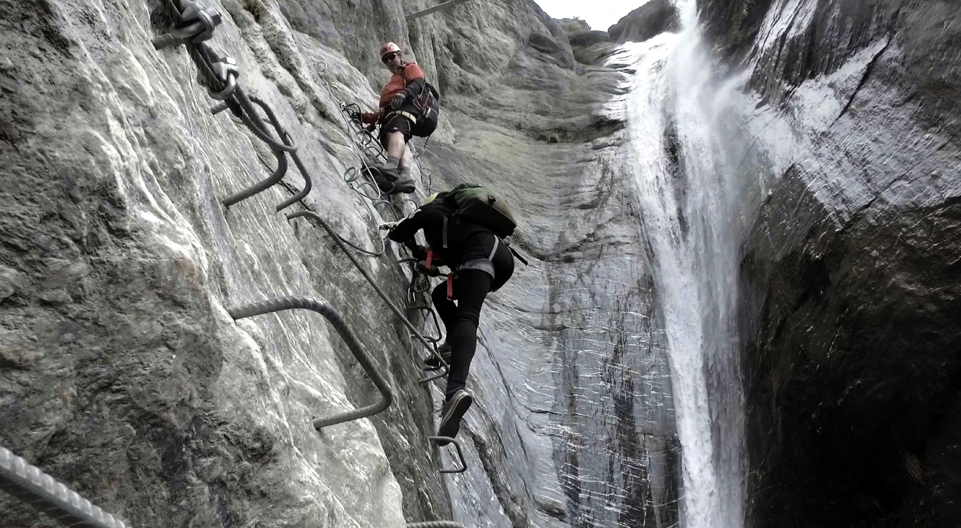 Two people climb up metal staples alongside a waterfall in New Zealand