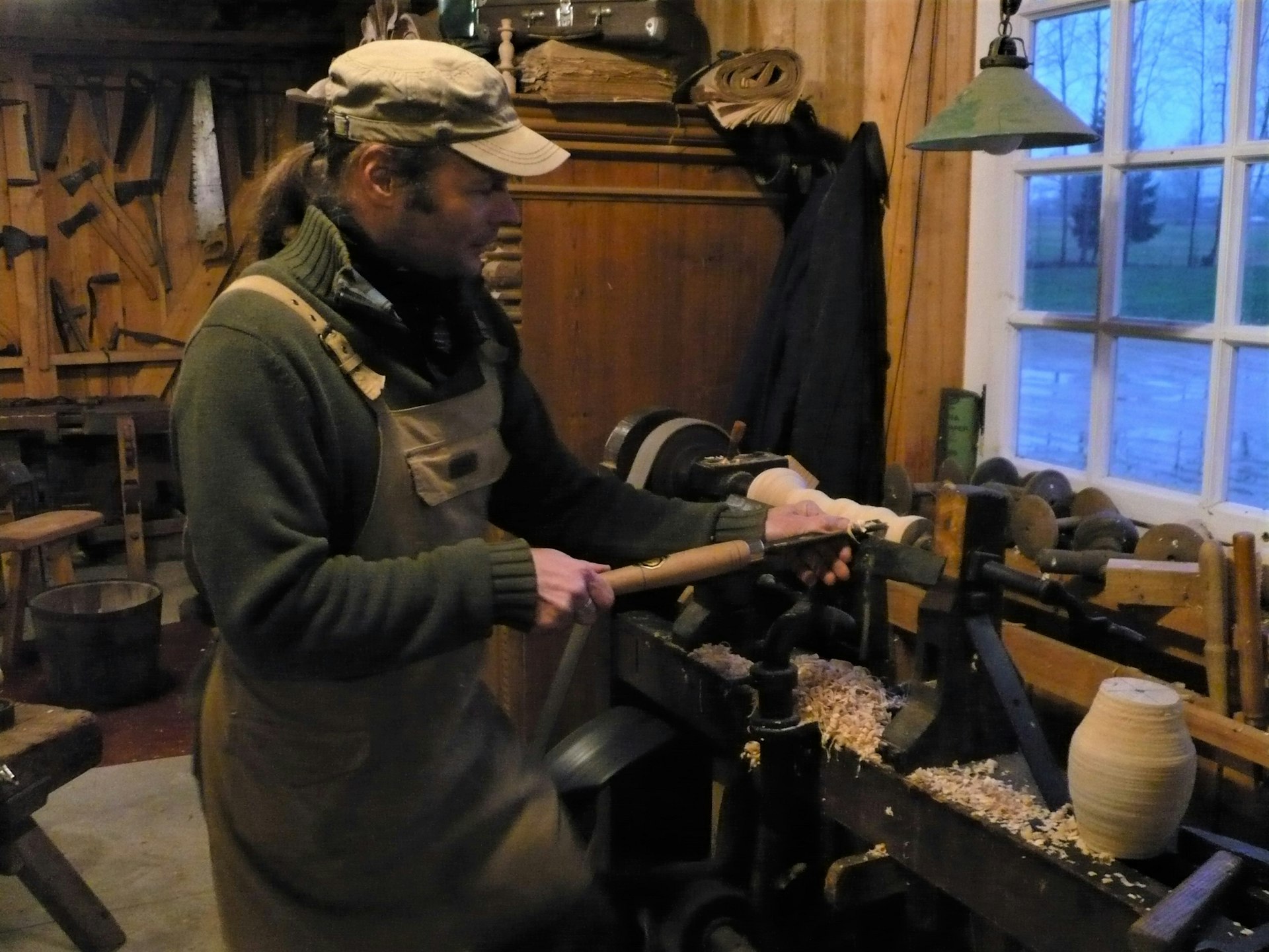 A man in a workshop leans over some wood in a vice. He's holding tools that are shaping the wood