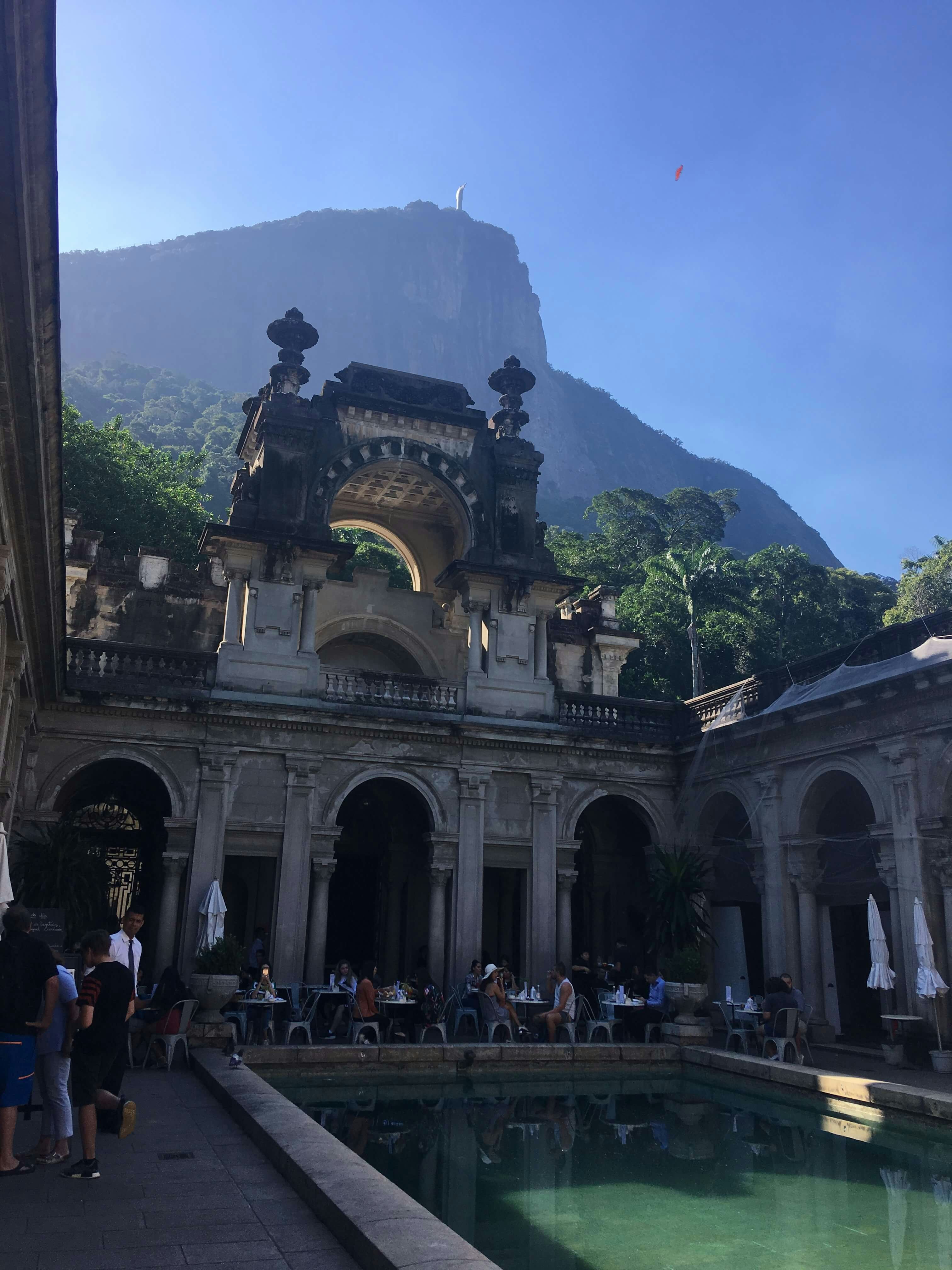 A small pool in the middle of a stone mansion with tables and chair. In the background, Corcovado mountain stands with a statue of Christ the Redeemer at the very top