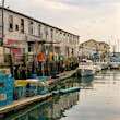 Wooden buildings and lobster traps line the Portland Pier near Luke's Lobster restaurant. Several small fishing boats float on the water as seagulls fly overhead