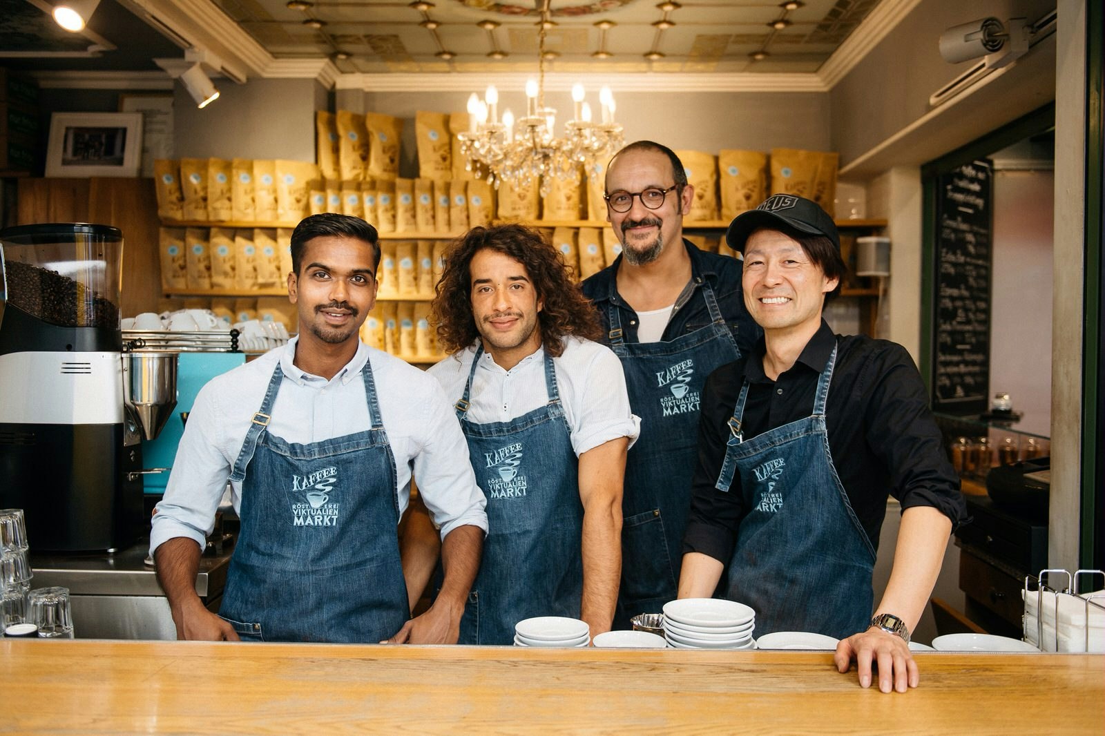Four men wearing denim aprons with the kaffeerosterei logo embroidered on them stand behind a wooden counter smiling at the camera. Behind them, out of focus, there are shelves stacked with bags of coffee beans and a chandelier hanging from an ornately-decorated ceiling. 