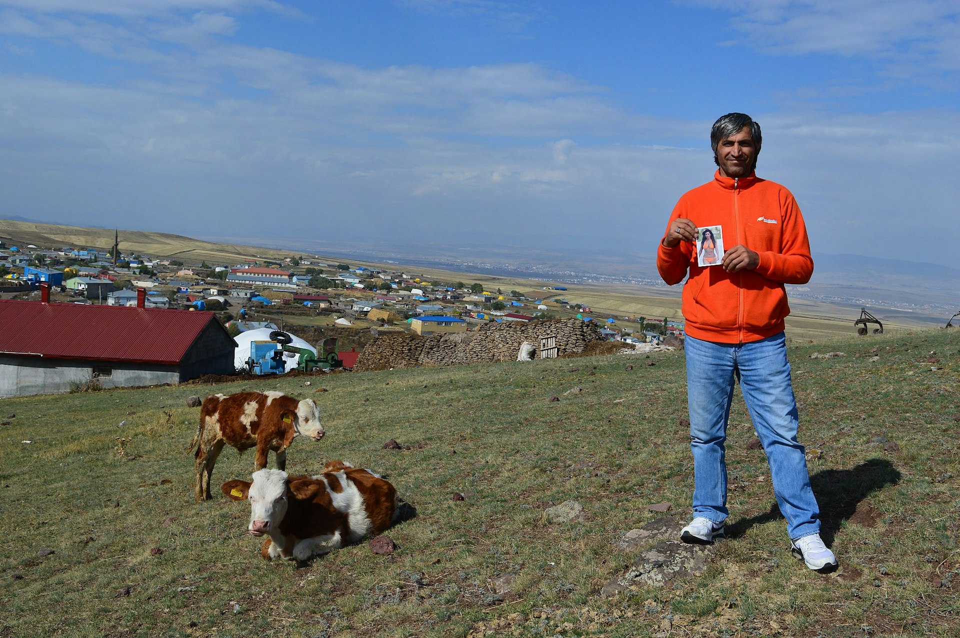 A villager wearing jeans and an orange fleece stand in a field holding a picture of Kim Kardashian; there are some brown and white cows next to him with the village in the background.