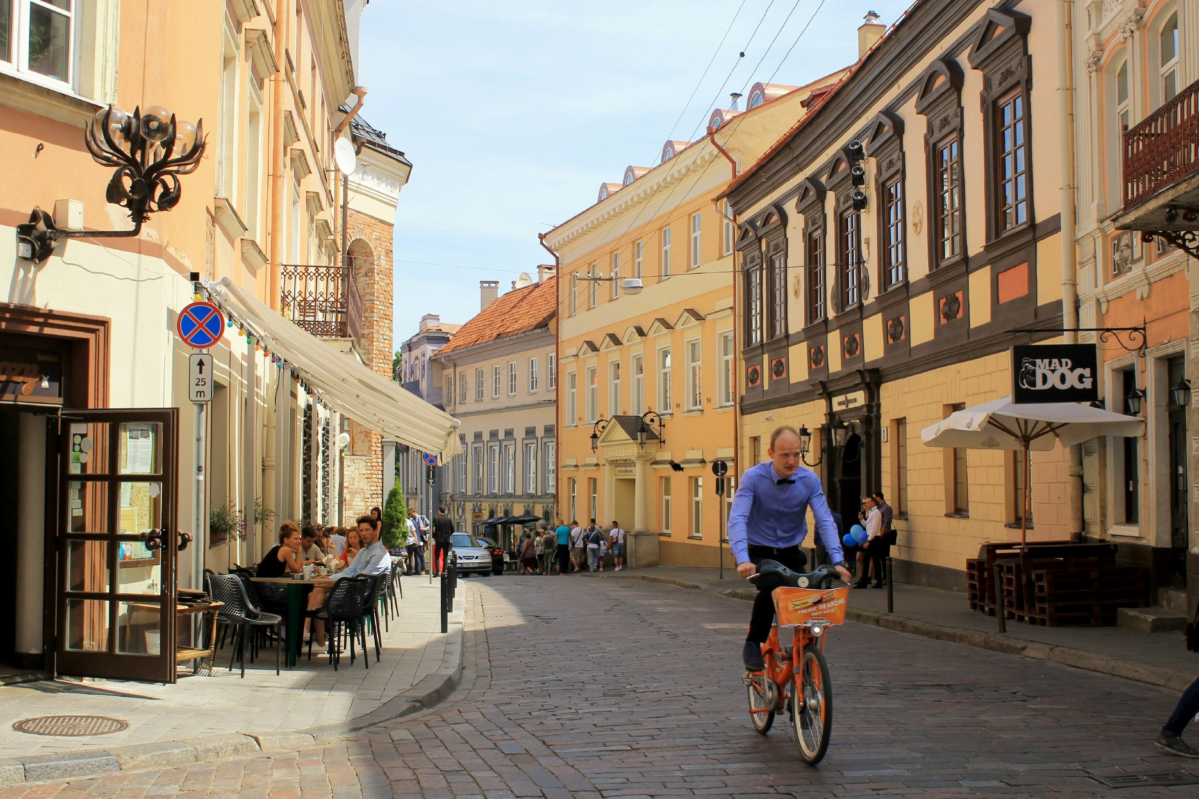 A man rides his bike through a street in Old Town Vilnius, as people sit at a cafe. 