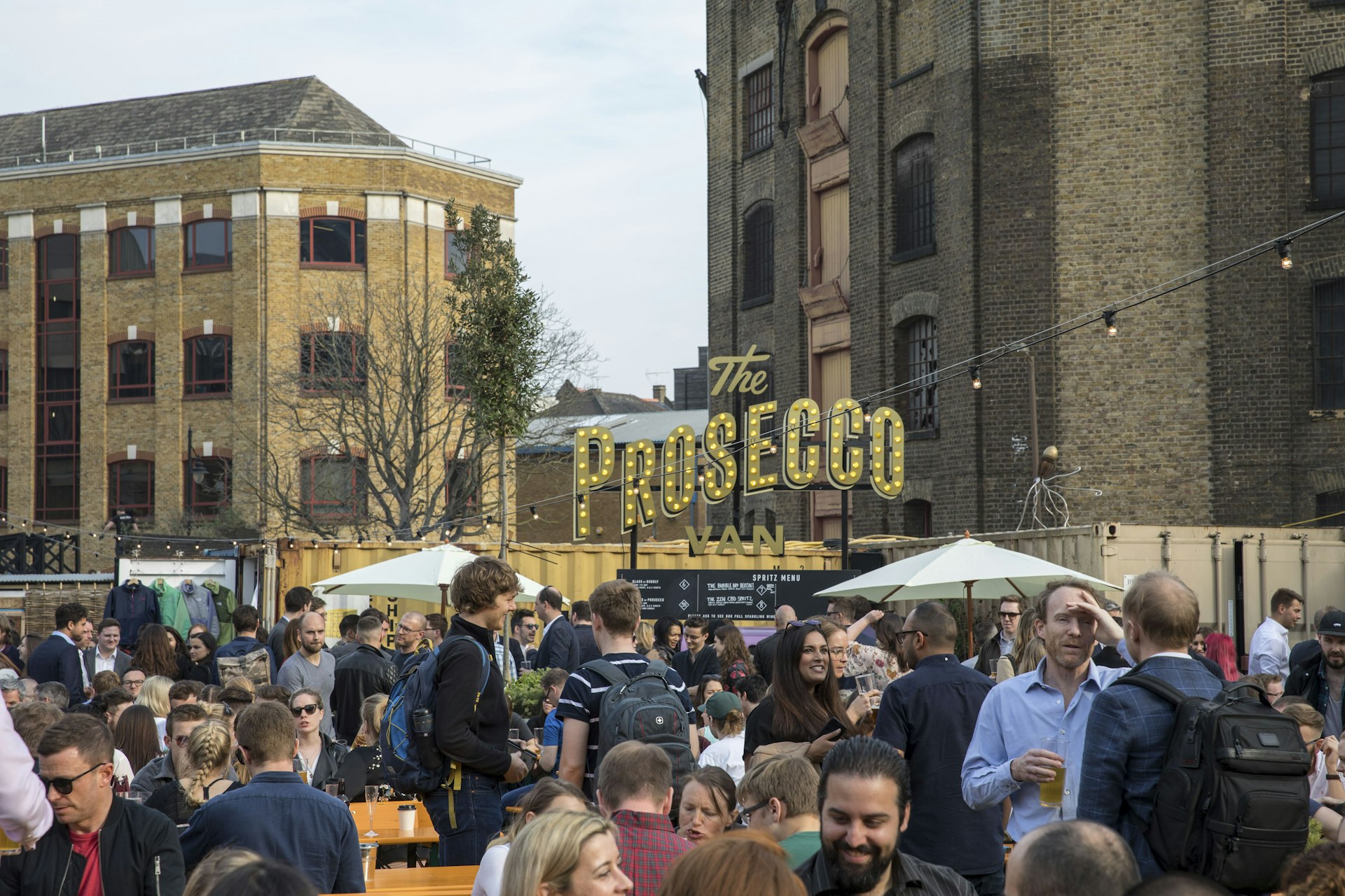 Industrial-looking brick buildings are the backdrop to a busy market scene, where people holding drinks mill between food stalls at London's Vinegar Yard market