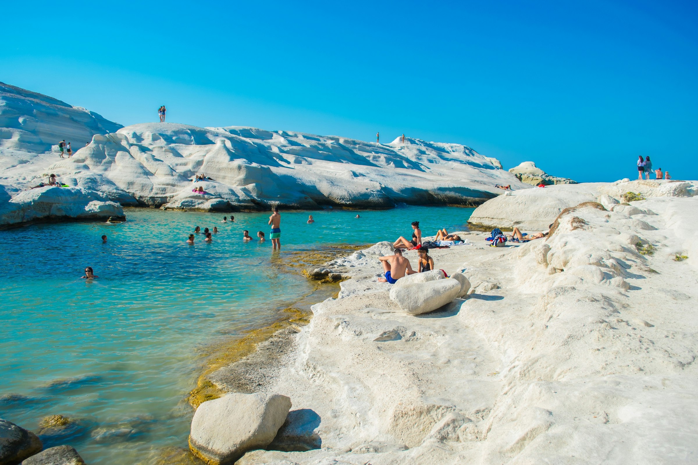 People sunbathing and playing on the white volcanic rocks that form the beach in Milos. The channel through the middle is turquoise and has about ten swimmers in it