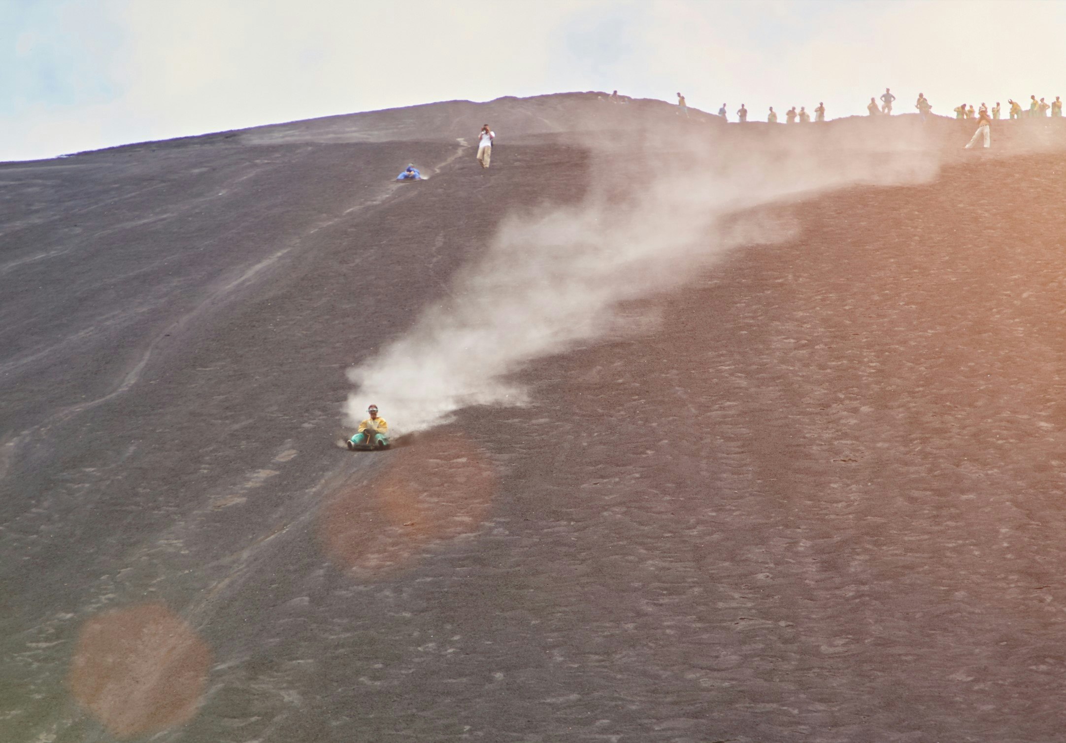 A man is sliding the side of a steep hill, on a volcano, while sat on a board. 