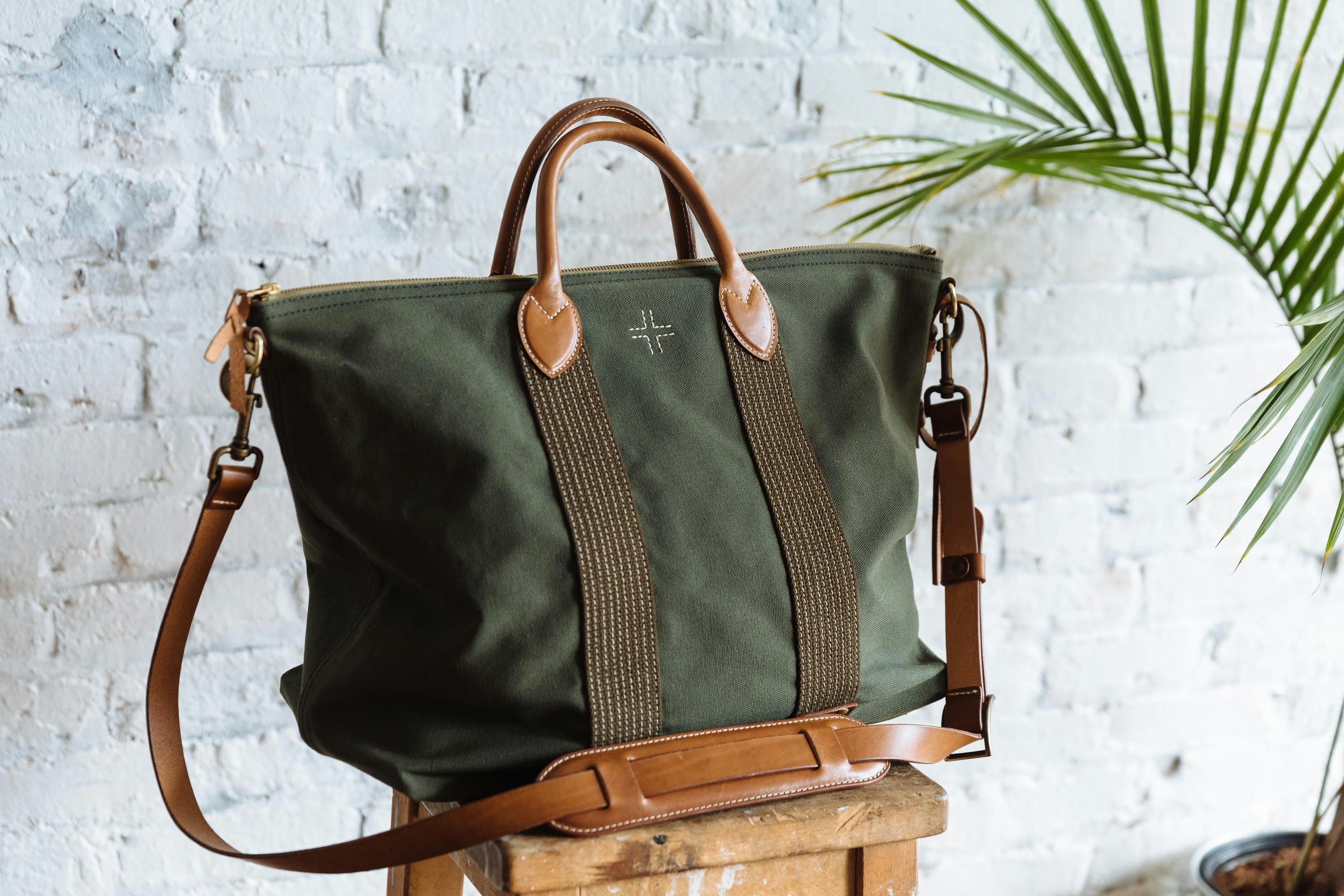 WP Standard's green canvas travel bag, with brown leather handles, a leather shoulder strap, and brown stripes on the front, sitting on a stool in front of a white brick wall and next to a palm frond