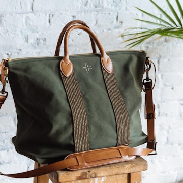WP Standard's green canvas travel bag, with brown leather handles, a leather shoulder strap, and brown stripes on the front, sitting on a stool in front of a white brick wall and next to a palm frond