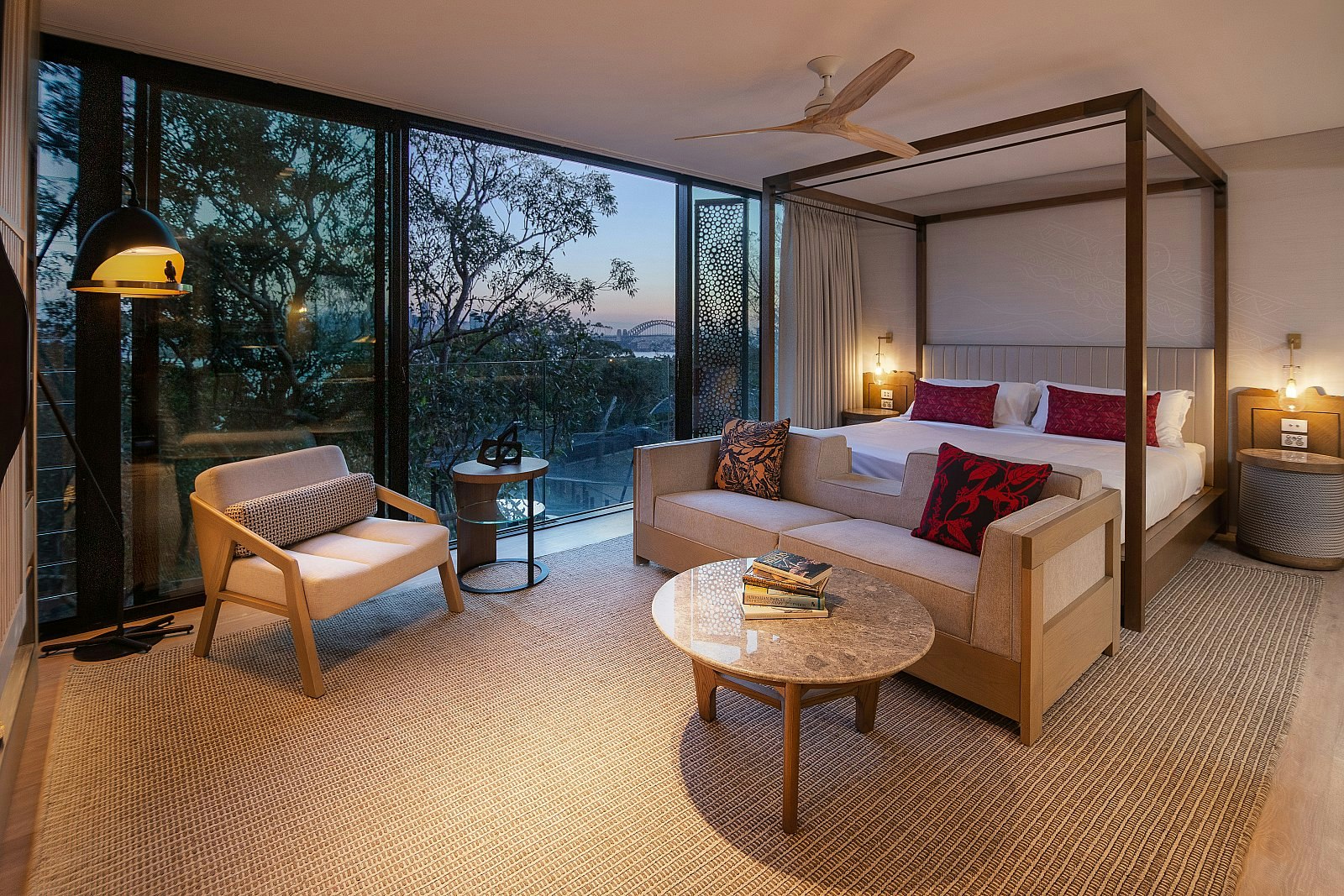 A large suite at the Taronga Zoo Wildlife Retreat with a four poster bed and modern furnishings; the window looks over trees with Sydney Harbour Bridge visible in the distance.