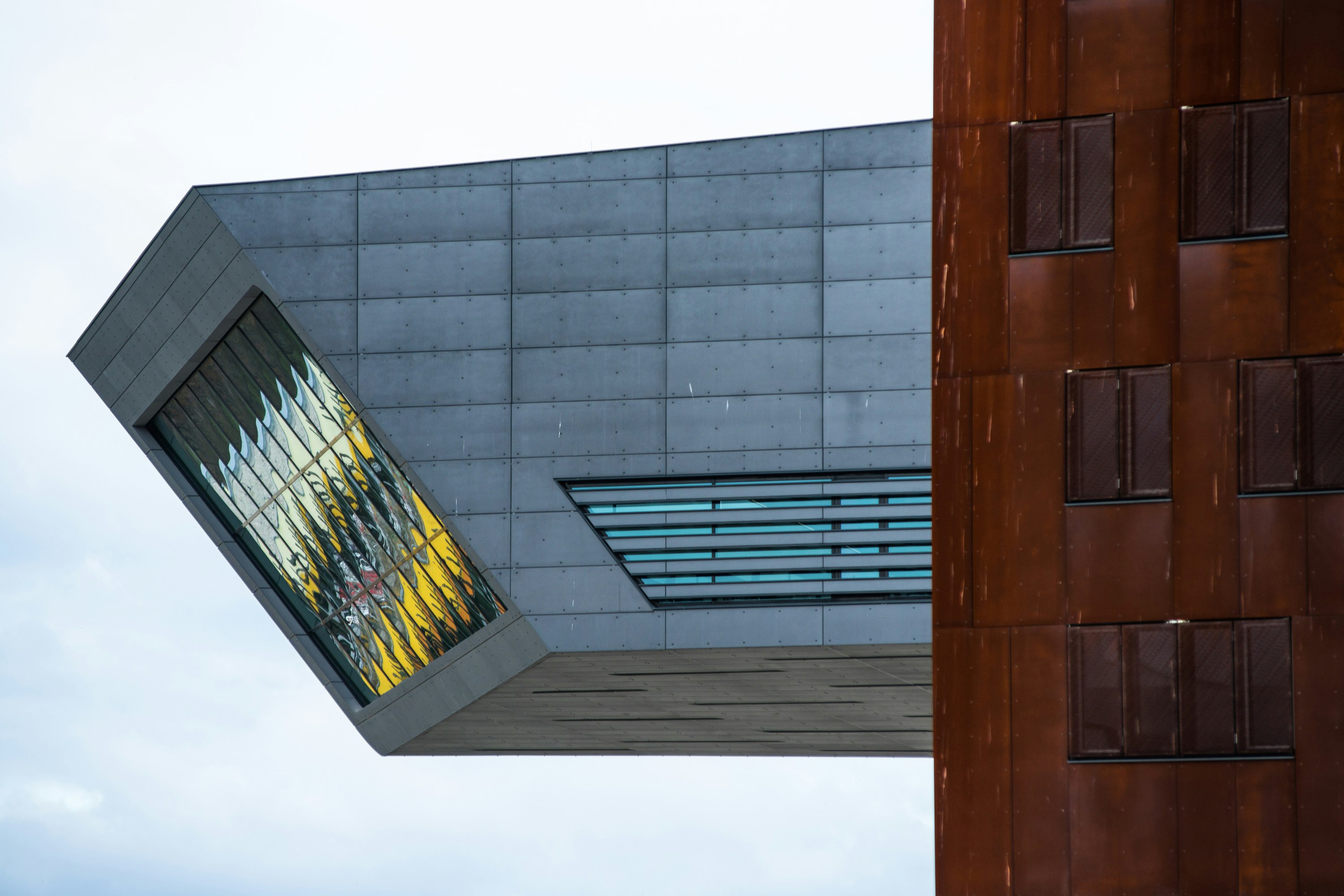A grey modernist cantilevered wing of the library at the Vienna University of Economics hangs from a brown structure and ends in with a rectangular glass wall of windows at the end, angled down towards the ground below, reflecting colors ranging from light blue to yellow