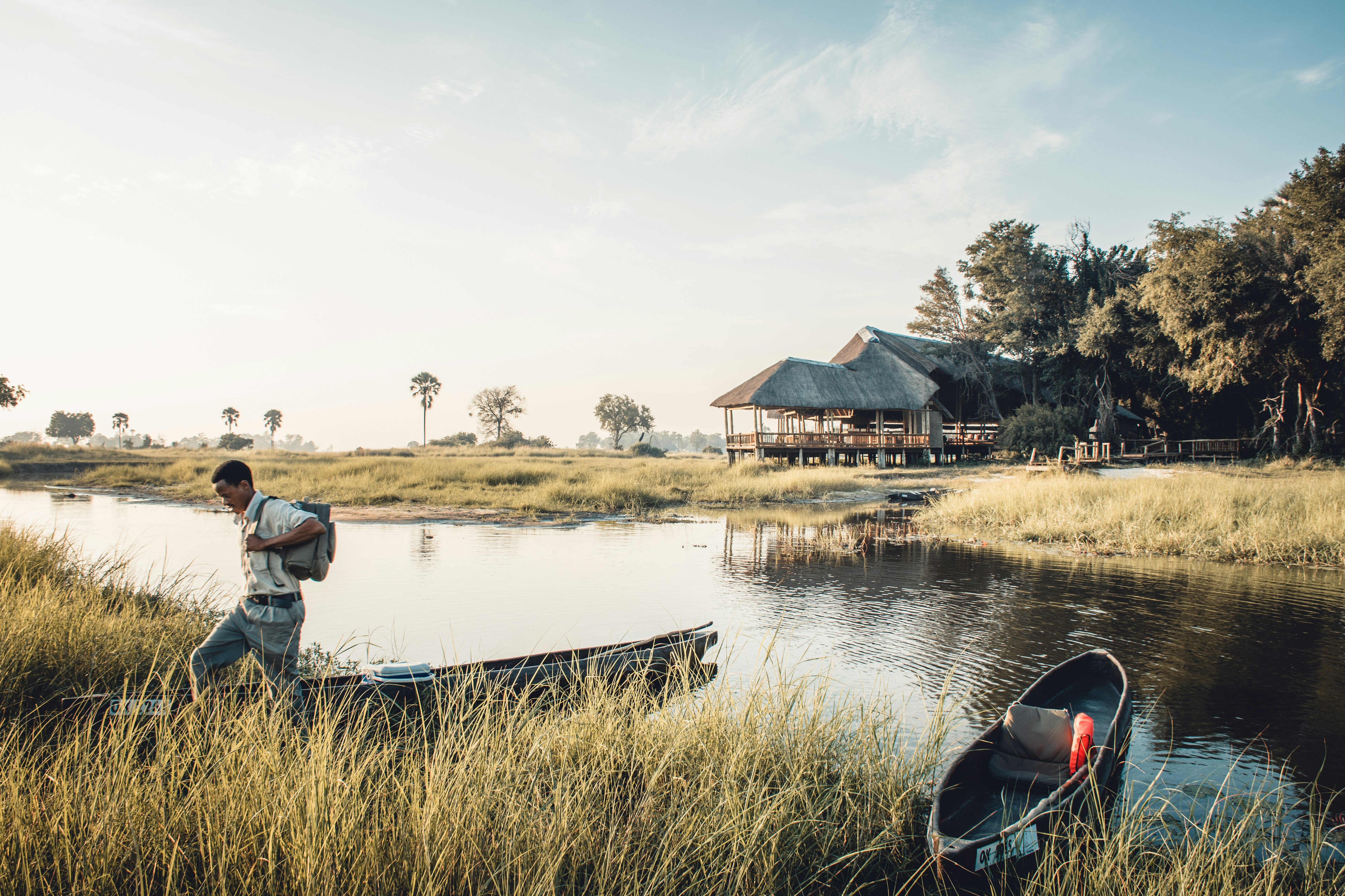 A man in grey twill slacks and a cream button down shirt with a full backpack steps through grasses on teh edge of the water where canoes are beached in the Okavango Delta. Across the water on the opposite shore are buildings with grass roofs and a cluster of trees.