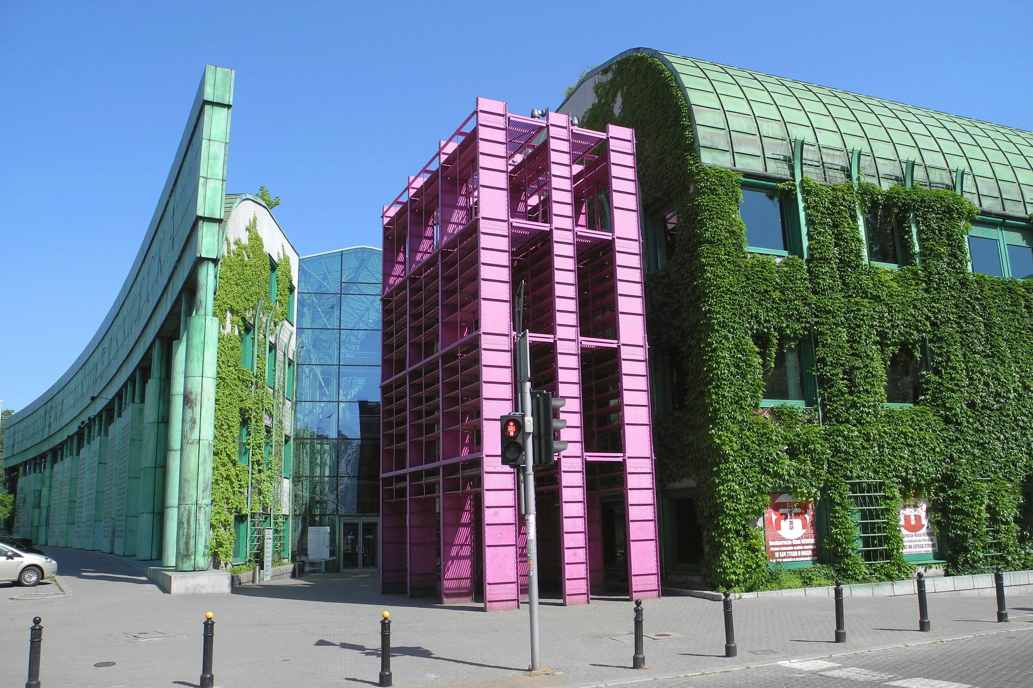 A gren curved wall made up of several panels meets a grey curved section covered in ivy, which in turn abuts a blue glass atrium. In front of the whole building is a pink section that looks almost like scaffolding. 