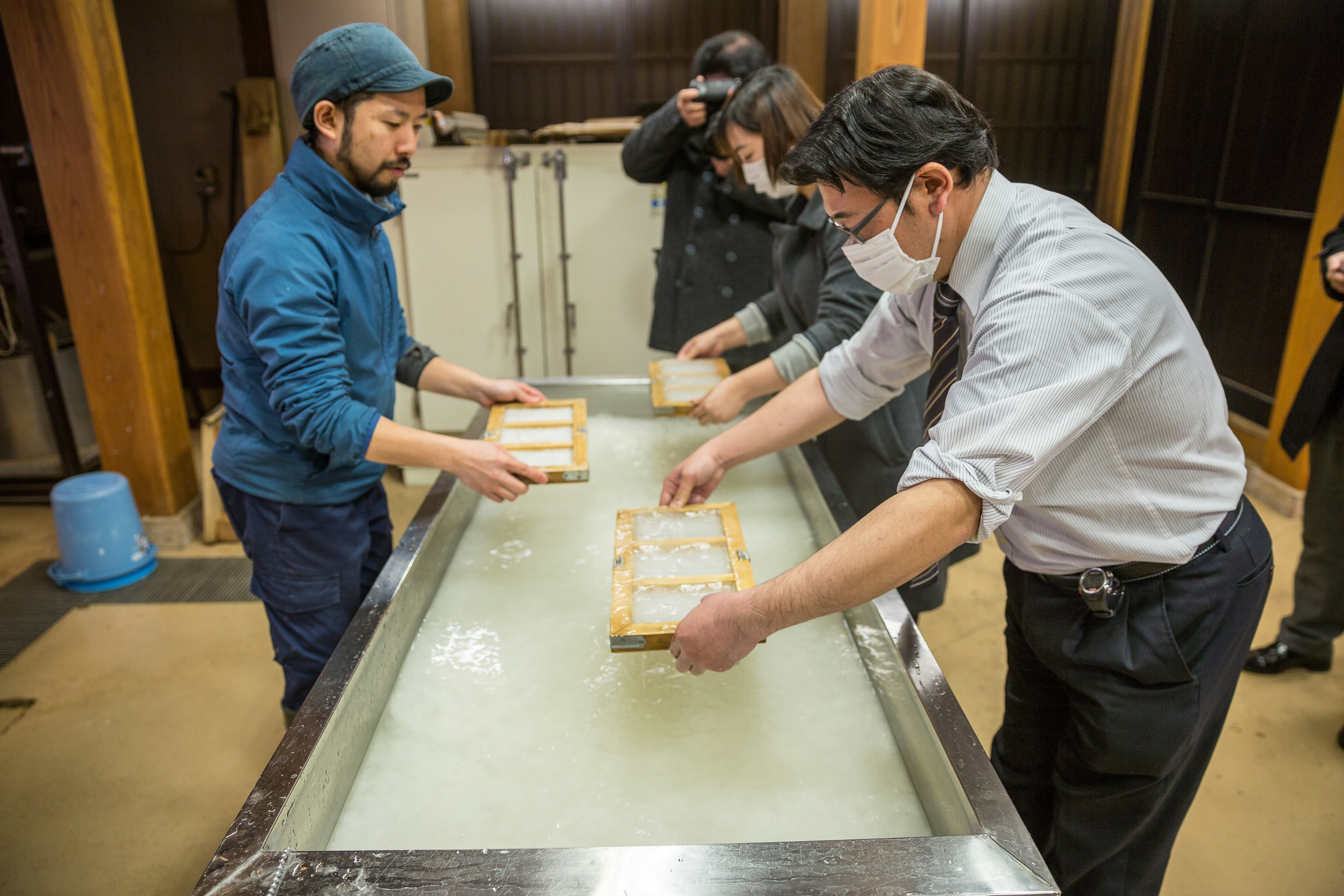 Three people lean over a trough of white liquid holding small wooden frames, which are the traditional tools used in making washi paper