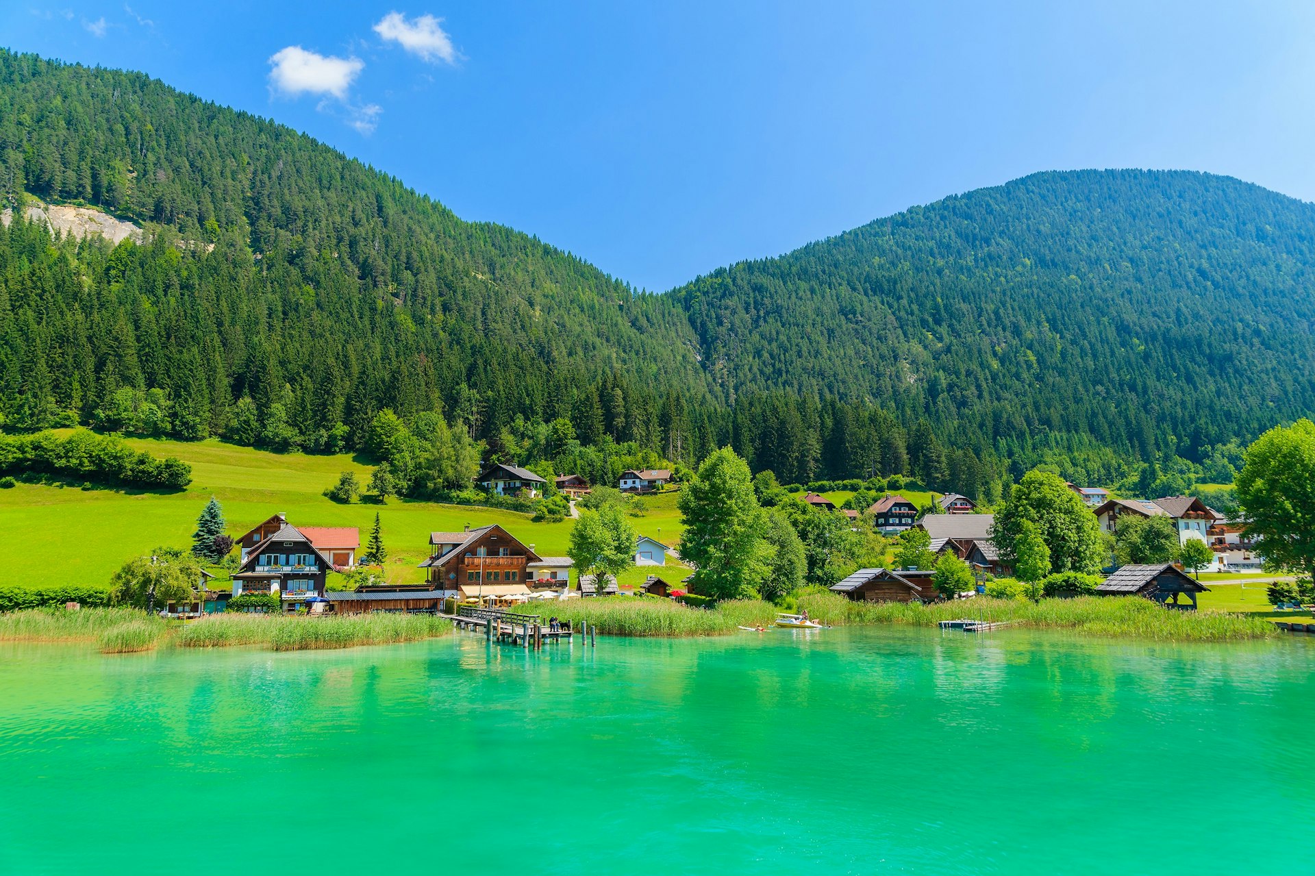 A bright green lake surrounded by hills covered in woodland. There are several chalet-style houses on the lakeside