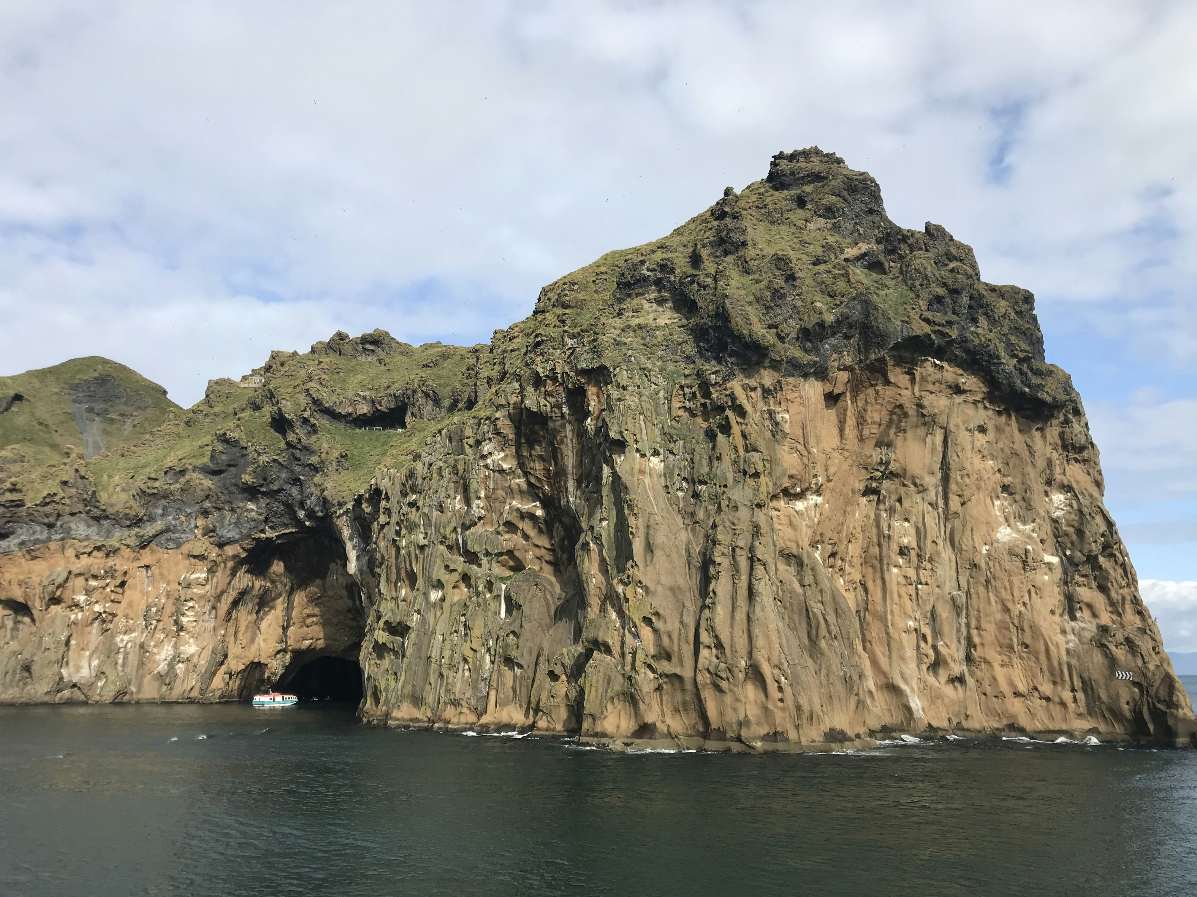 The brown, craggy cliffs of Westman Islands are full of black pockets where puffins nest and streaked with small white patches of guano. The islands are topped with green on the very top layer of the rock, and are surrounded by deep blueish green water. A small blue and white boat sales into a cave or inlet in the left of the frame.