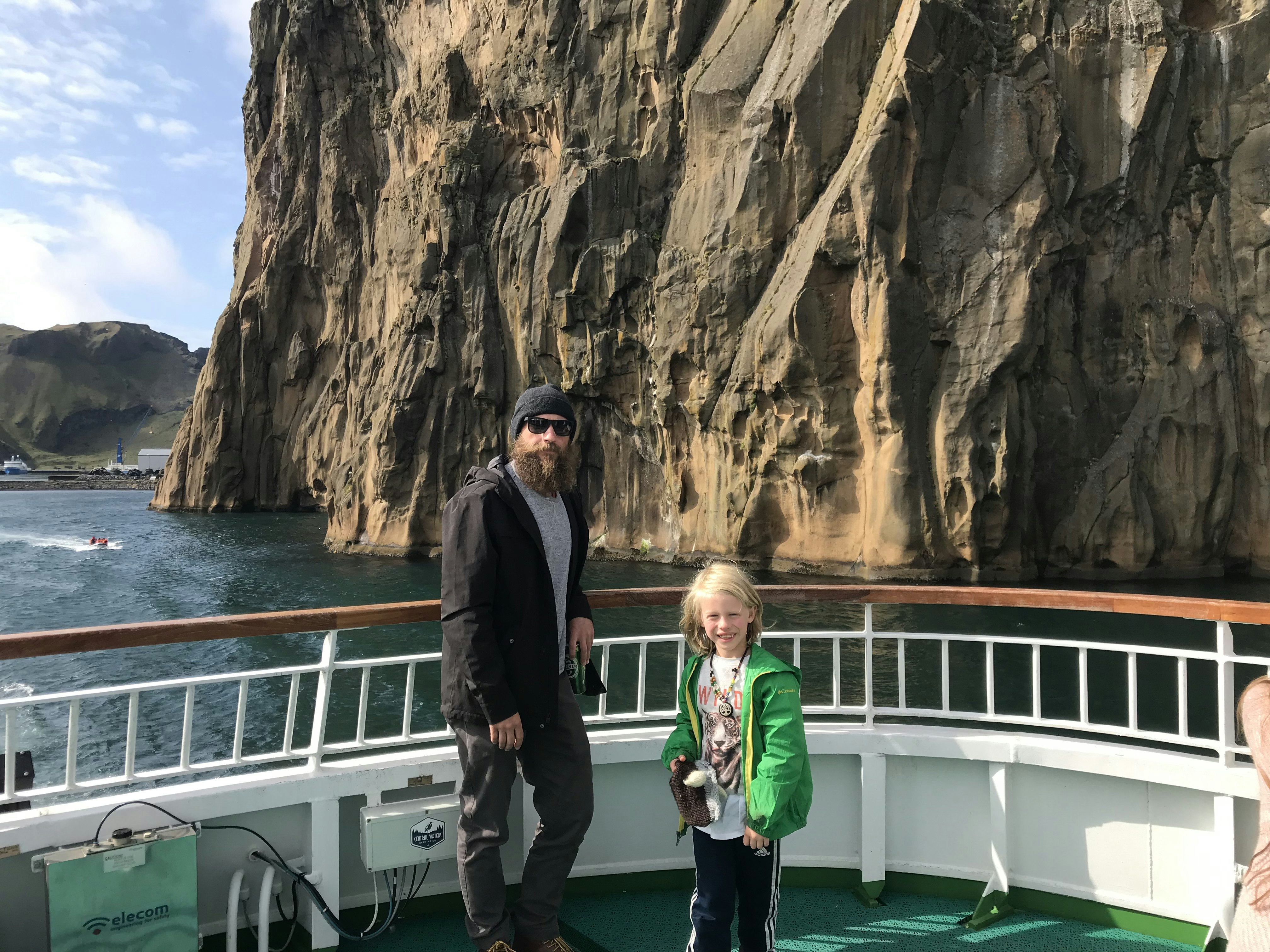 A man in grey slacks, a black windbreaker, and a heathered grey T-shirt, with a big bushy beard and black wayfarers and a fleece black watch cap stands on the bow of the Westman Islands Ferry with a small, tow-headed boy with a tiger on his t-shirt. The cliff face is in the background.