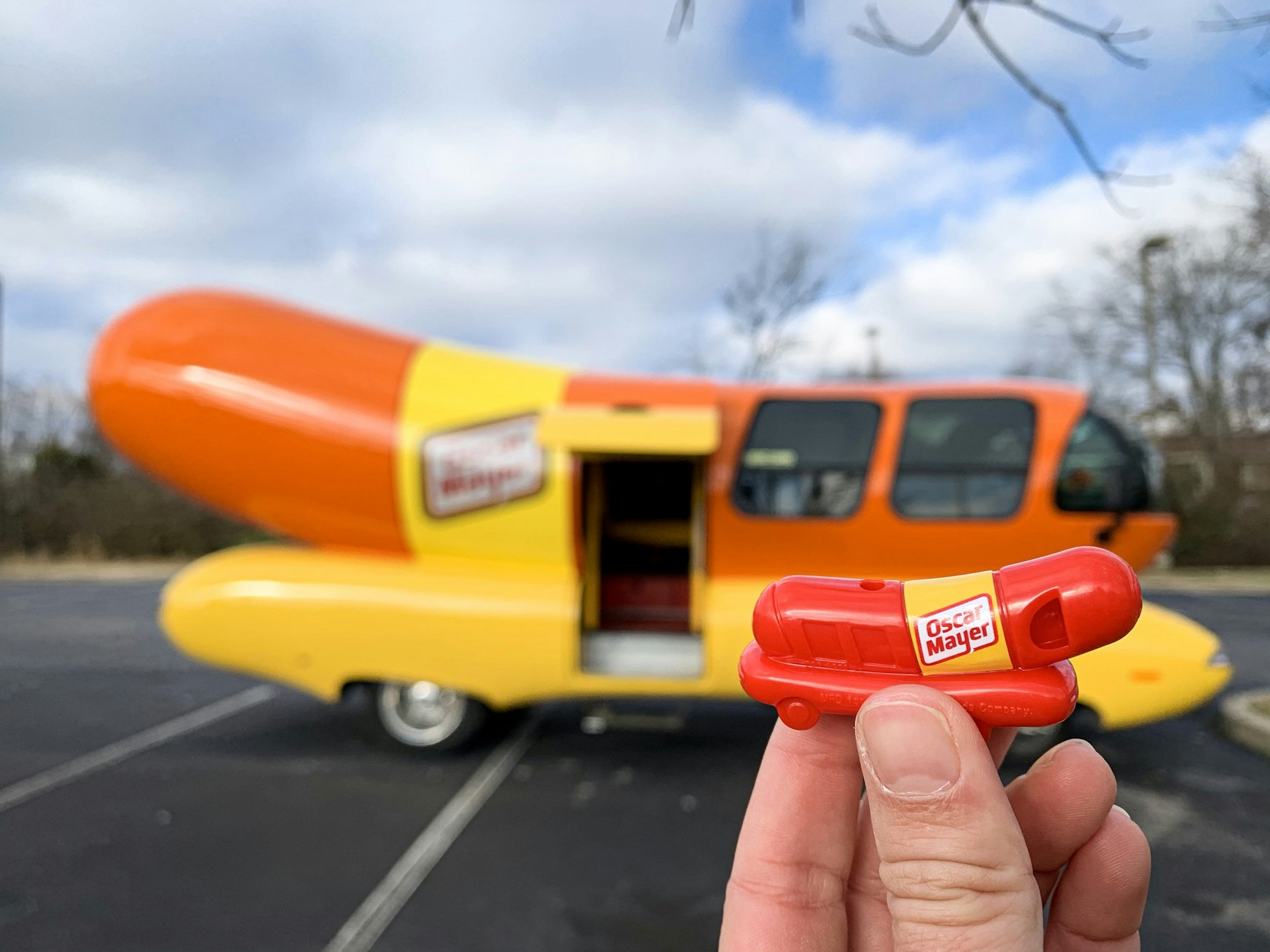 En hand håller i en leksak Weenie Whistle med Oscar Mayer Wienermobile i bakgrunden
