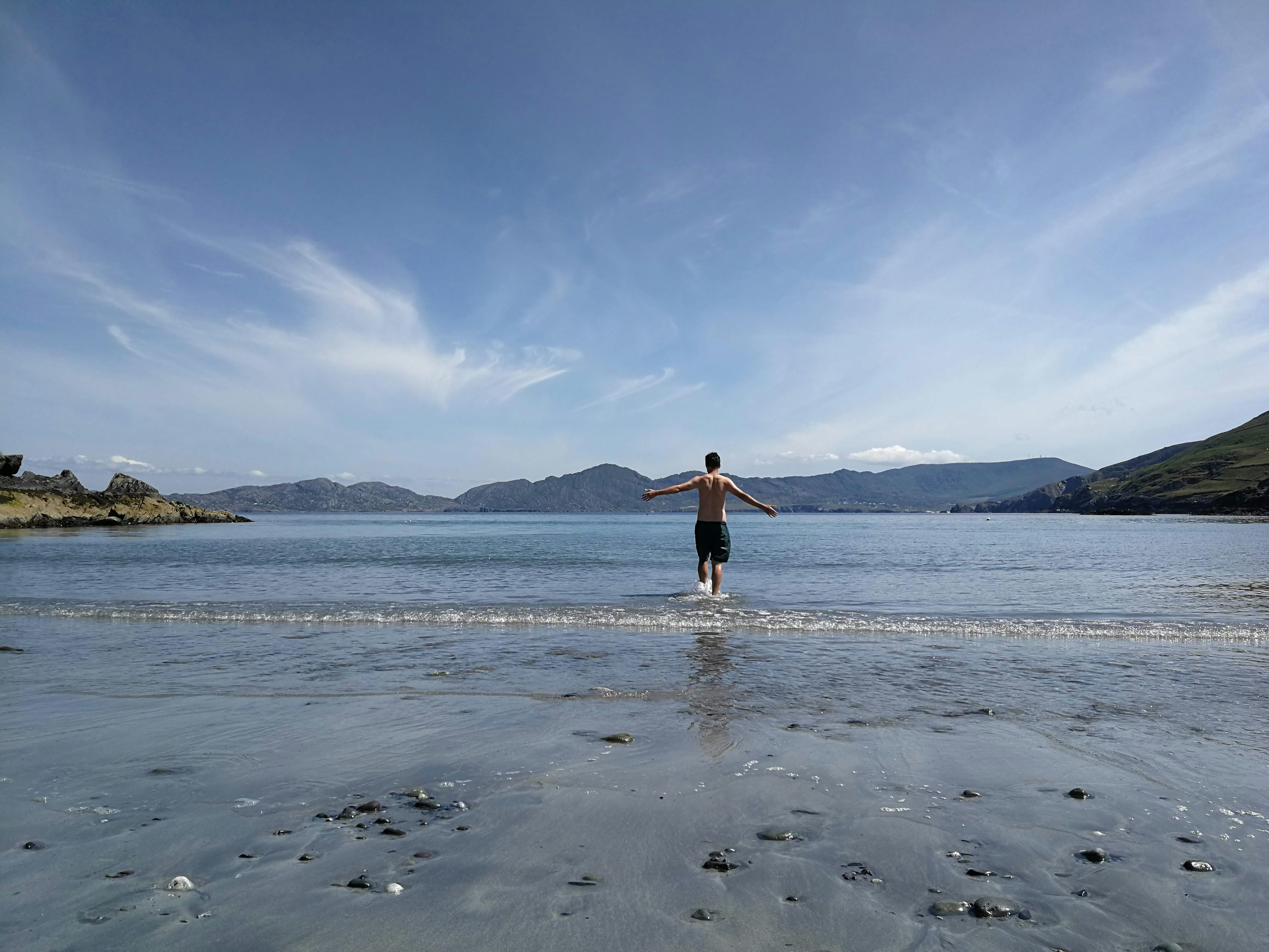 A rugged beach scene; the sky is bright blue and has wispy clouds, and craggy hills can be seen in the distance. Writer Joe Davis, wearing swimming shorts, is walking out into the calm water, his arms outstretched.