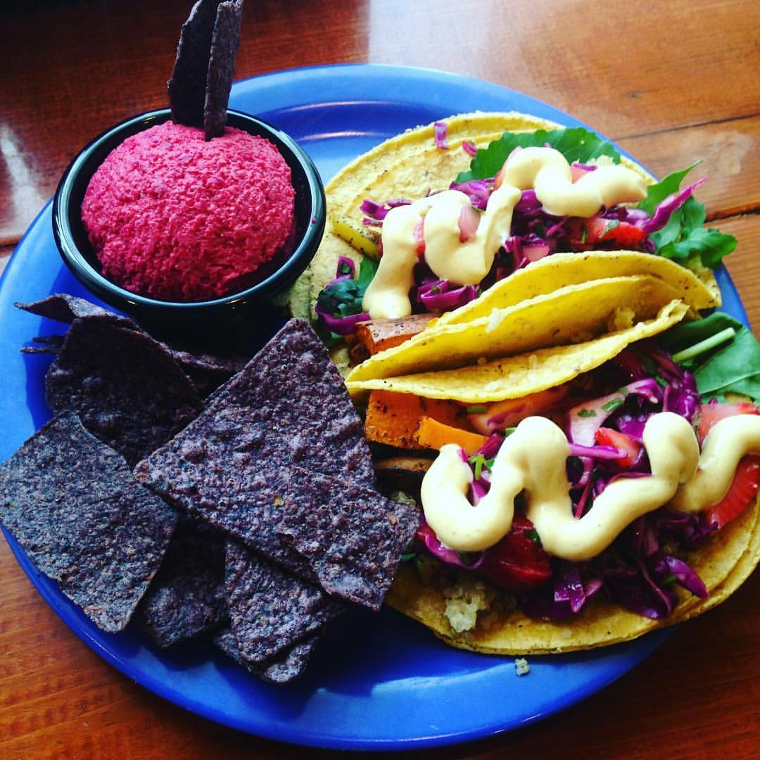 Electric blue plate holding a pair of soft-shell tacos filled with vegetables and herbs and topped with a creamy sauce; blue tortilla chips and small bowl filled with bright pink sauce.