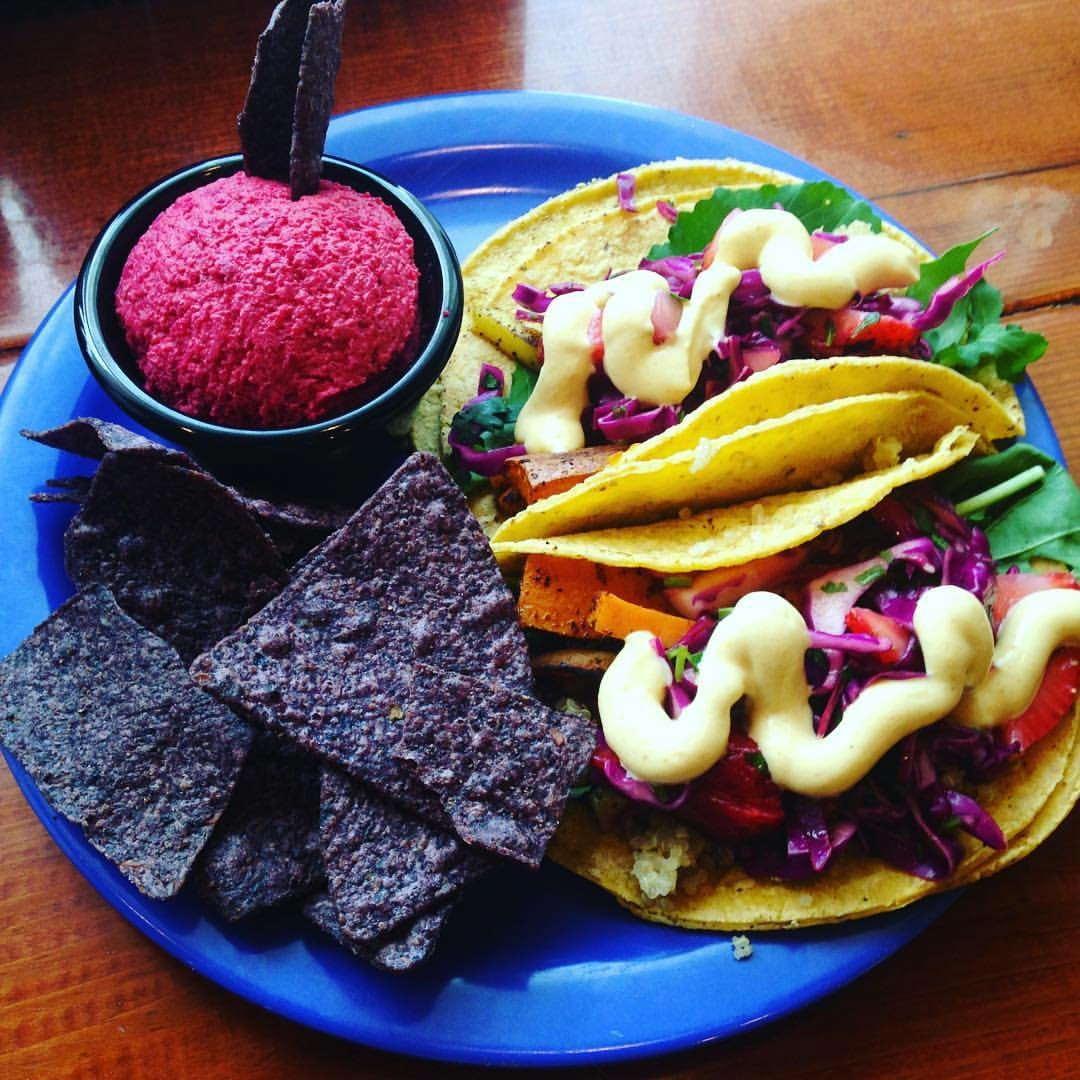 Elektrisk blå tallrik som rymmer ett par soft-shell tacos fyllda med grönsaker och örter och toppad med en krämig sås;  blå tortillachips och liten skål fylld med ljusrosa sås.