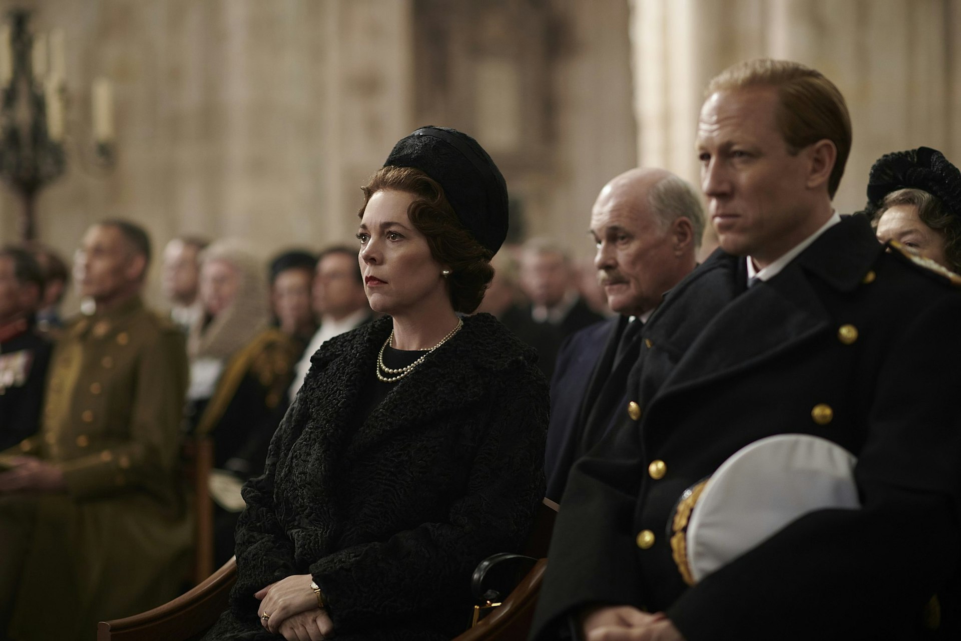 A still from The Crown season 3; Olivia Colman as the Queen and Tobias Menzies as Prince Philip are dressed in black at a cathedral funeral service.