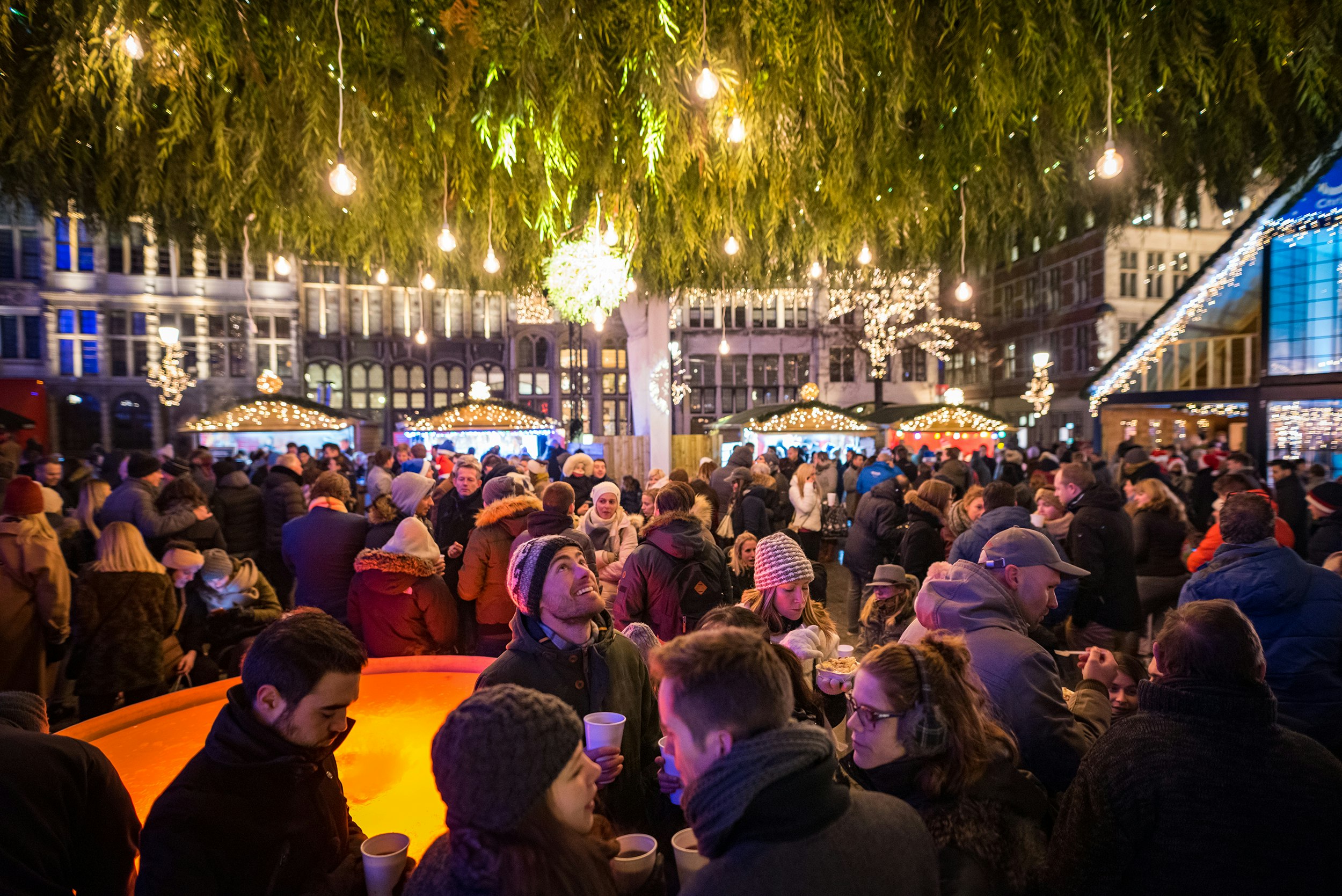 People drinking warm beverages at night under a canopy of lights and greenery surrounded by a Christmas market