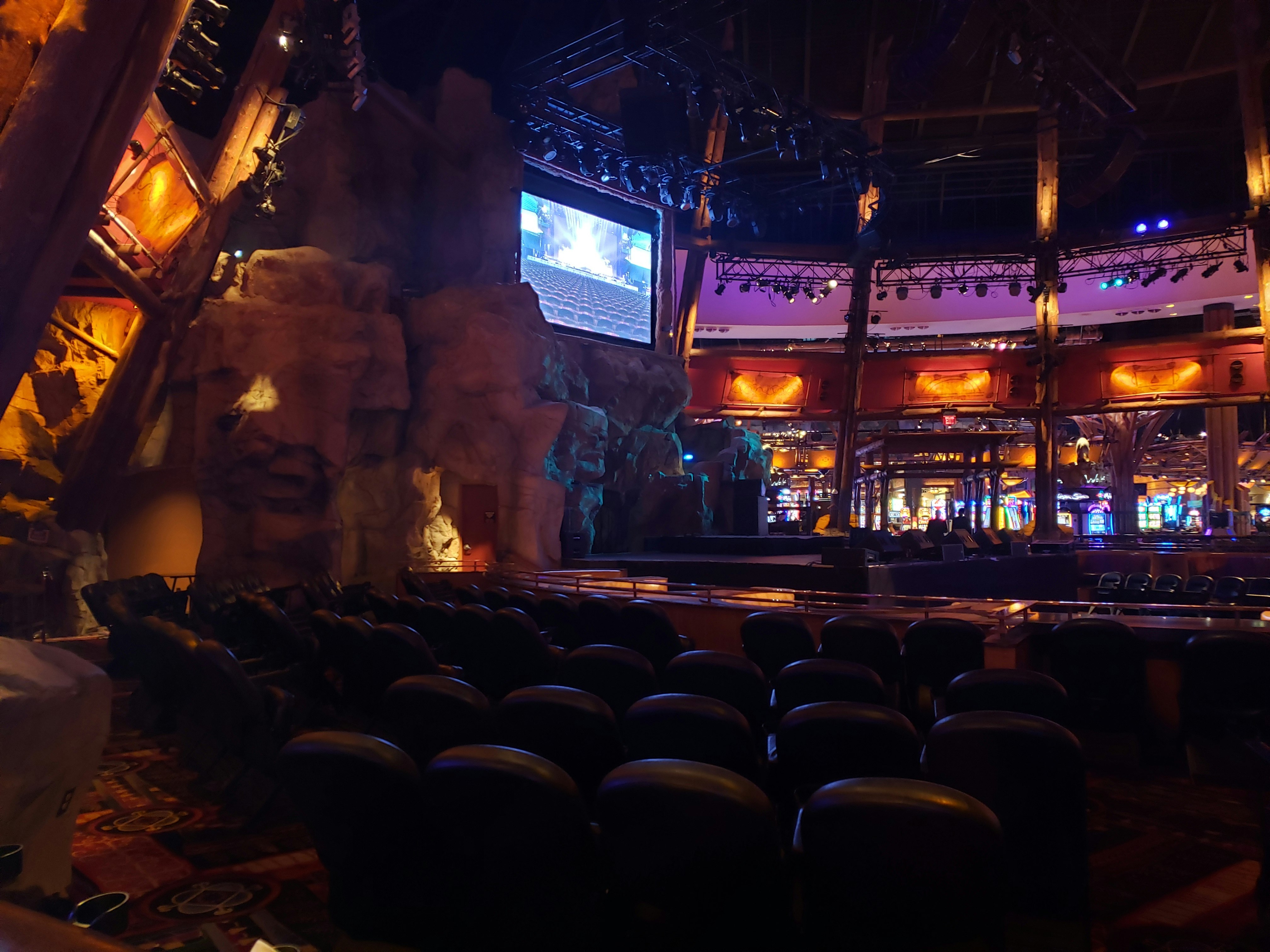 The Wolf Den at Mohegan Sun Casino has walls that look like giant boulders or a cave, with black vinyl seats with curved backrests in four rows in front of a large stage lit with purple and orange lights. A big flatscreen TV display over the stage is showing footage from another, larger amphitheater. 
