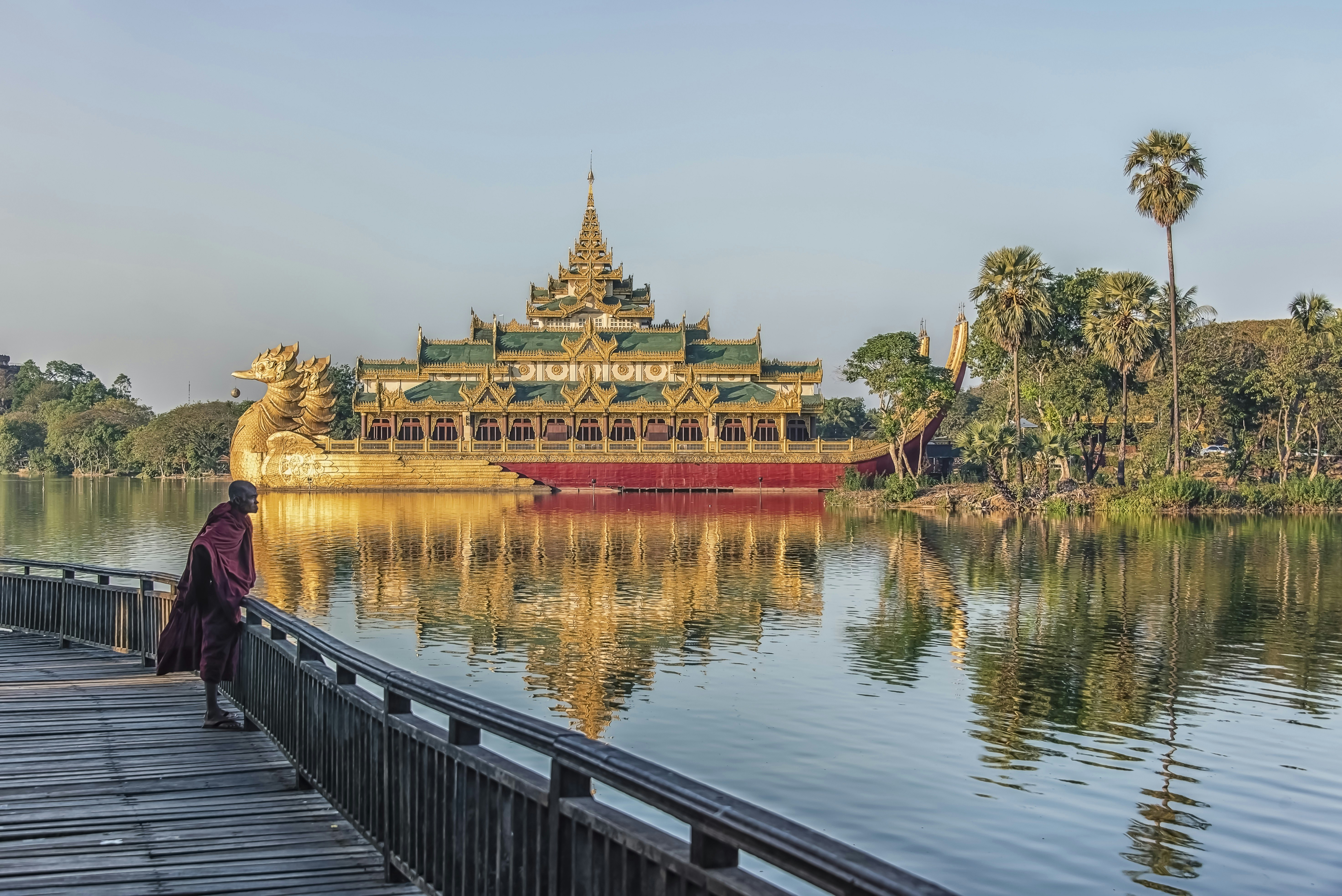 A monk, dressed in a red robe, stands on the boardwalk at Kandawgyi Lake looking out over the water. In the background, the Karaweik Palace looms large over the lake.