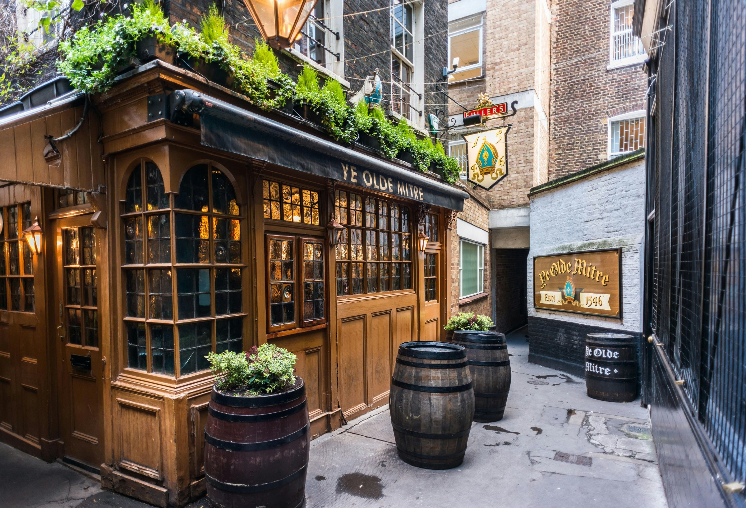 The wooden-fronted Ye Olde Mitre pub exterior with oak barrels outside as tables and green plants running above the awning