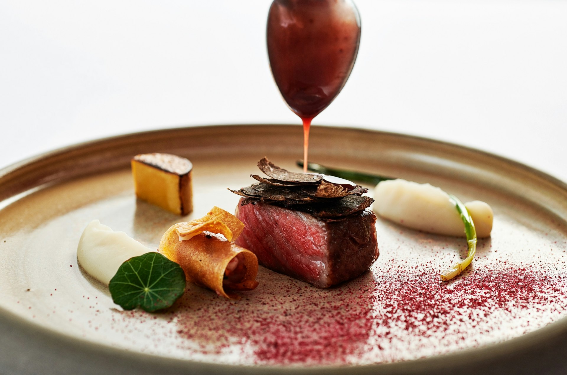 A spoon hovers over a small cube of red meat, with jus flowing down; next to the meat are delicate morsels of food (celeriac mash, rolled up crisp etc) and a sprinkle of purple dust.