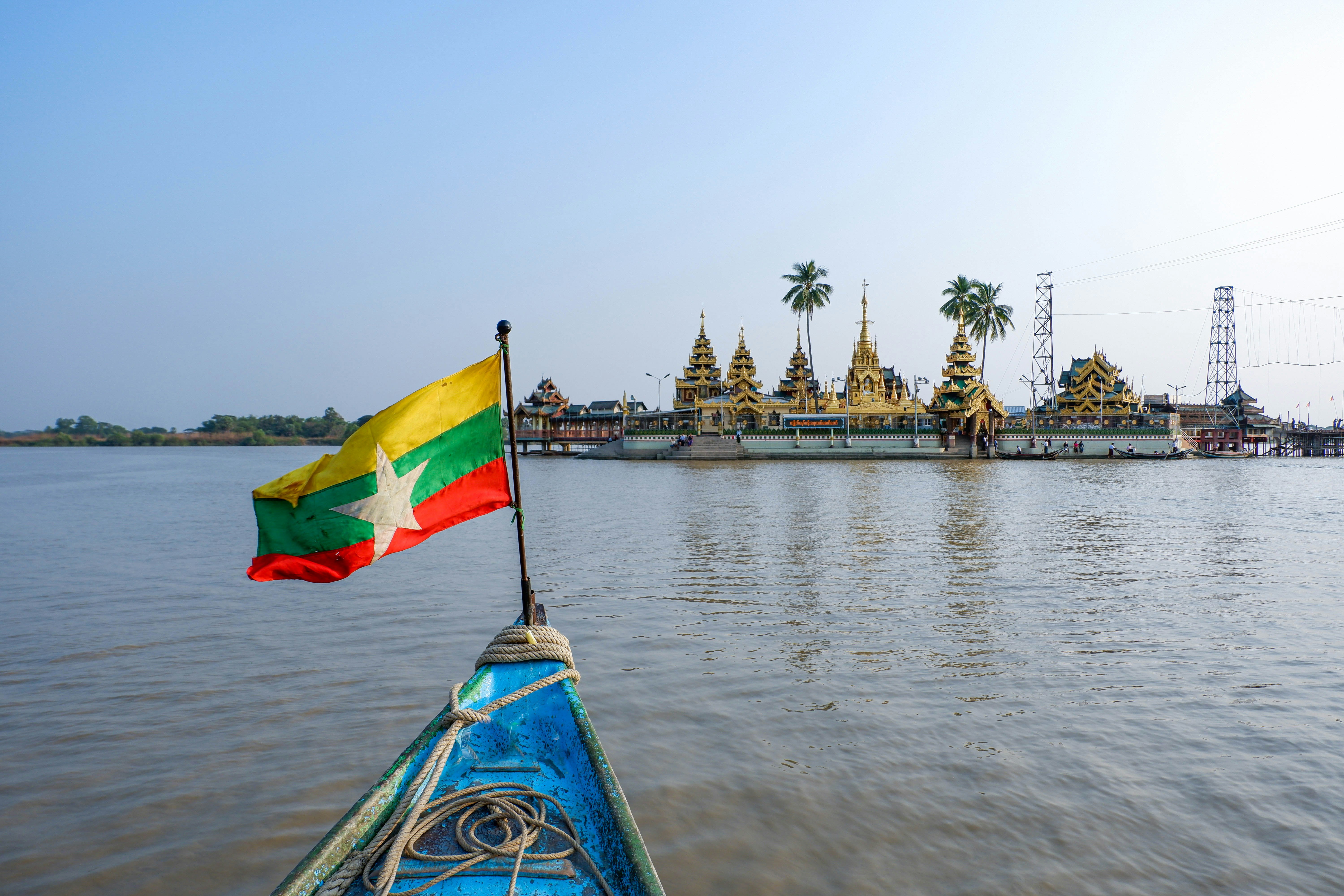 The front of a boat, flying the yellow, green and red flag of Myanmar, points in the direction of Yele river pagoda. A golden Buddhist pagoda built on a platform juts out into the river.