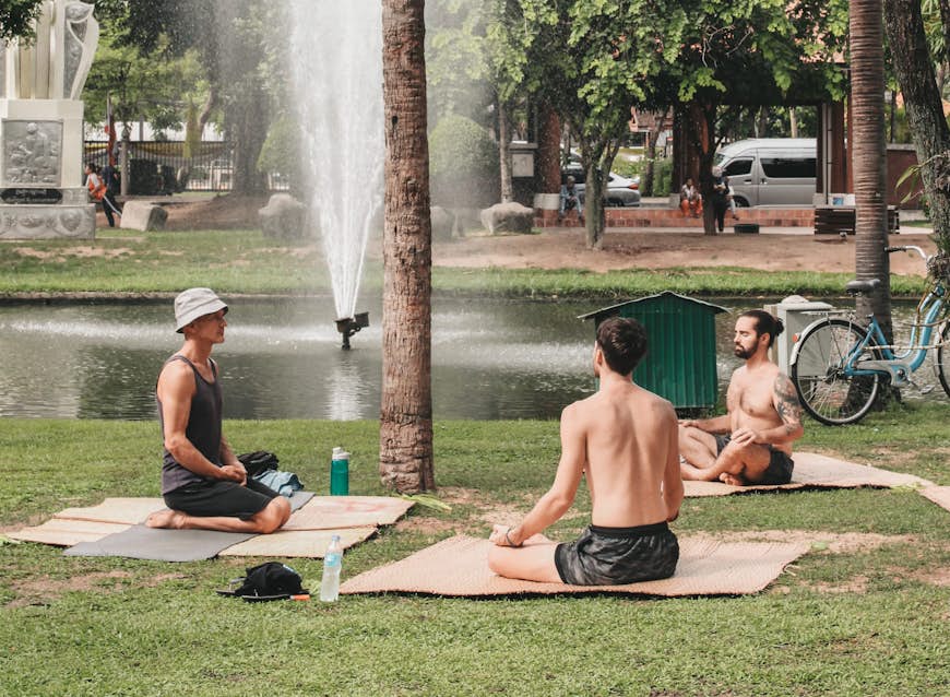 Three men sit on the grass in a Chiang Mai park practising yoga. In the background a small lake is visible with a fountain in the middle.