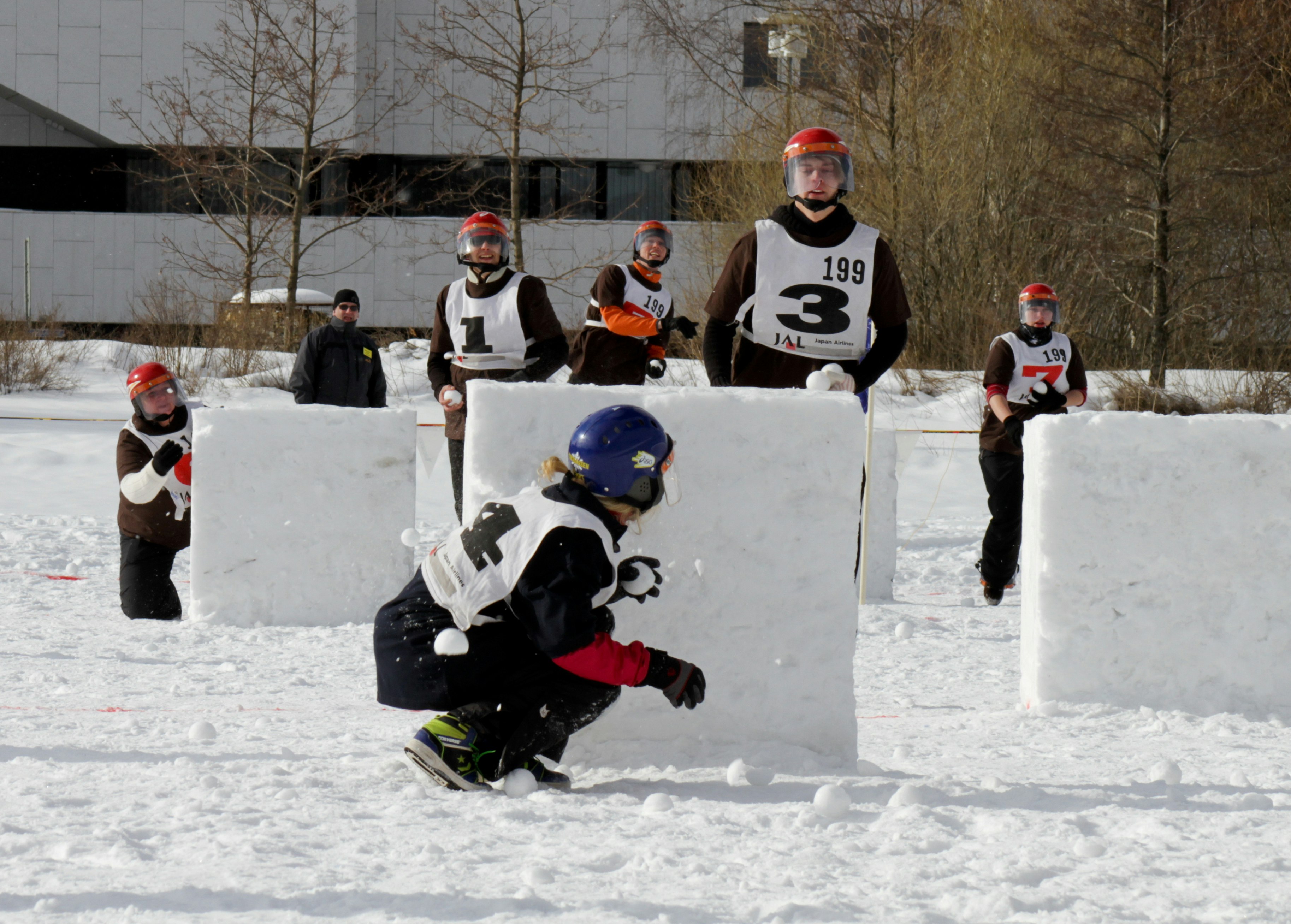 A person wearing a blue helmet and holding a snowball crotches behind a snow barrier as five people with red helmets advance in their direction during a snowball competition in Finland.