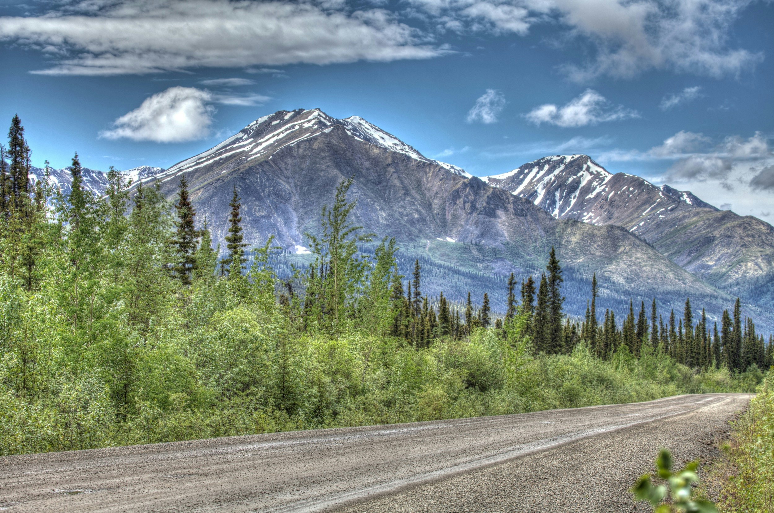 A rocky shale road - the Dempster Highway - runs through an evergreen forest at the base of a snow-covered mountain in the Yukon Territory