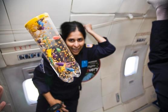 A clear plastic container filled with colorful objects, floating in the foreground in front of a Zero G researcher