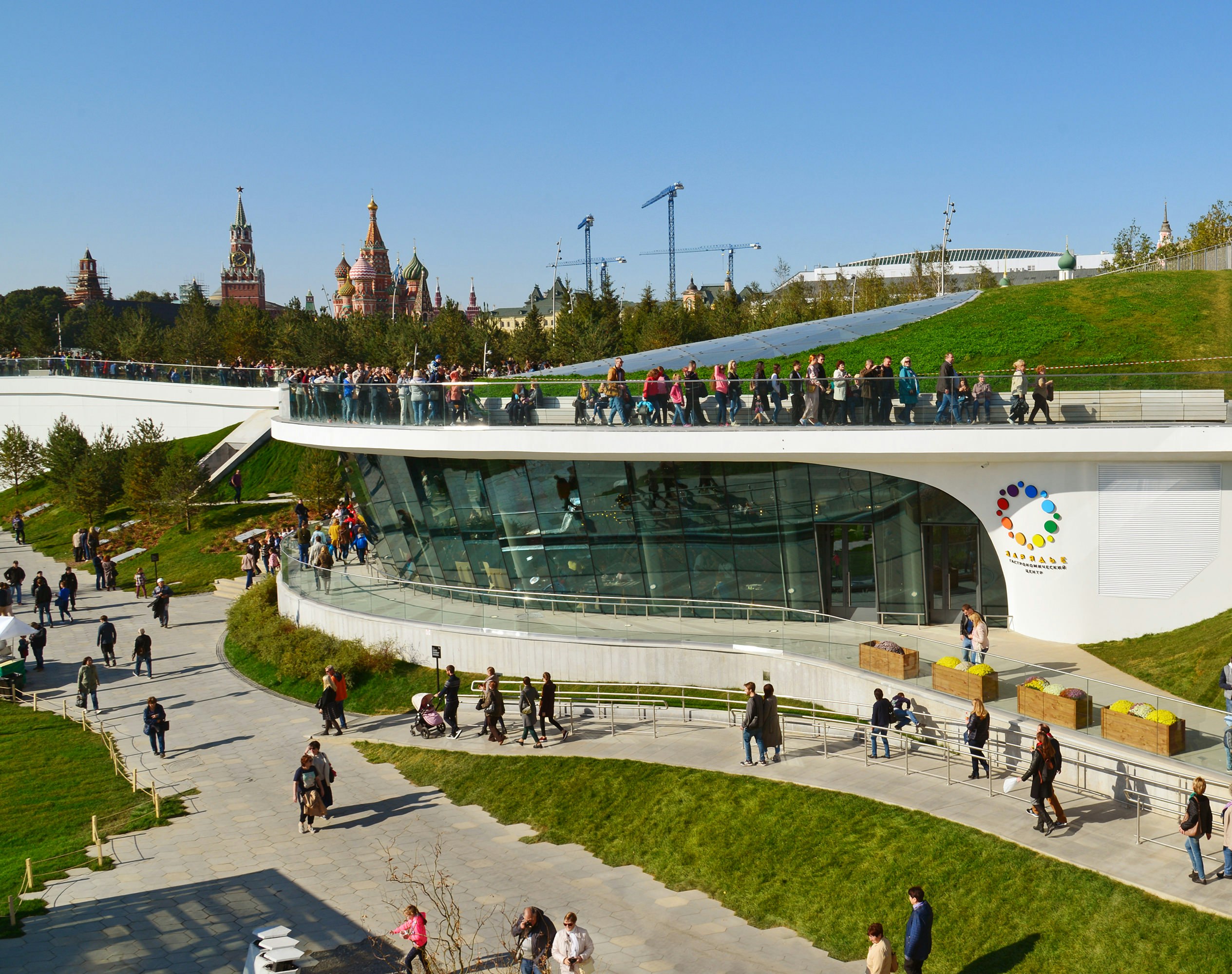 Zaryadye - a landscape and architectural park. Many people stroll over concrete walkways surrounding a rounded building in parkland.