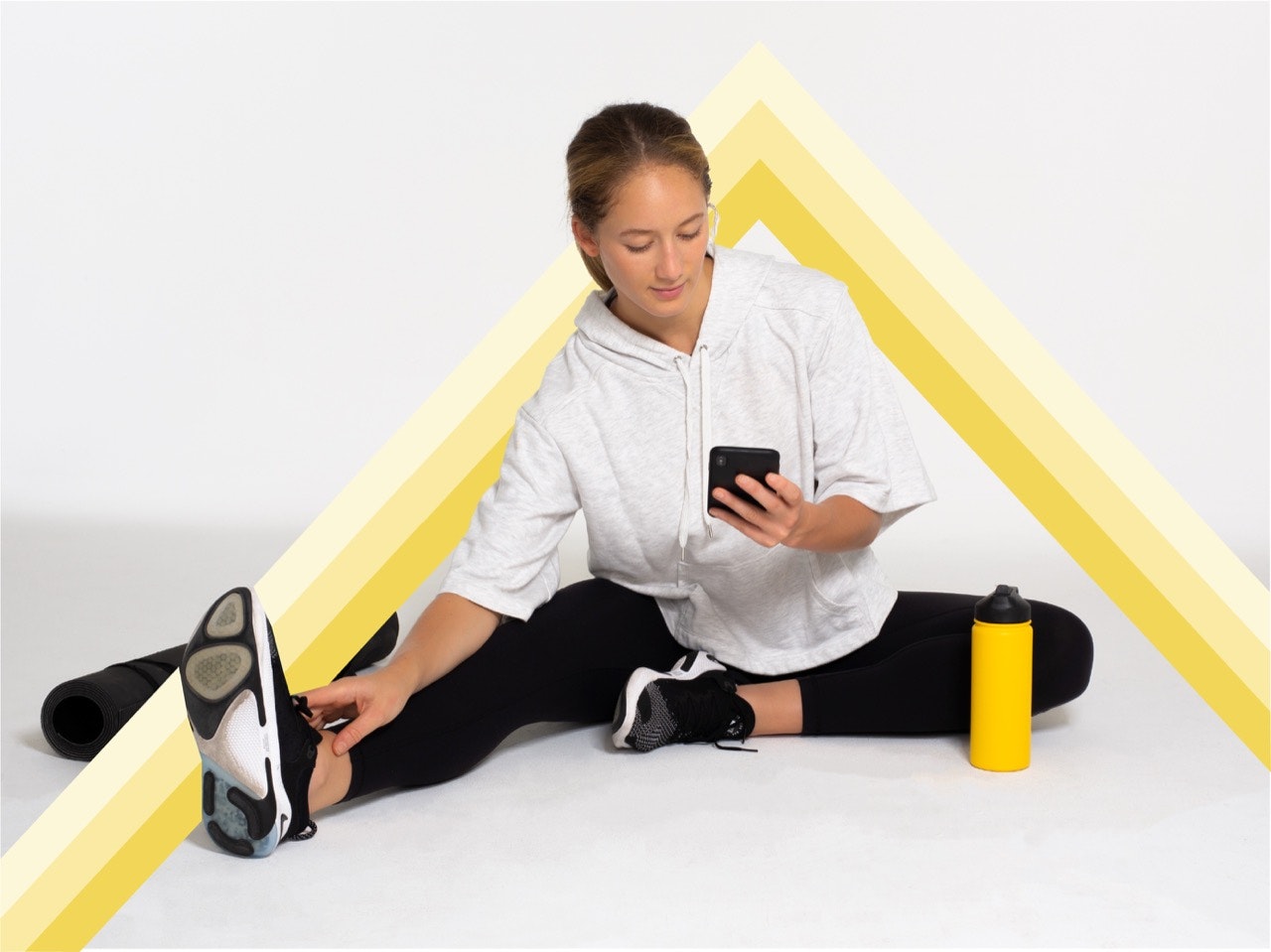 Woman sitting on the ground under a yellow triangle, stretching and looking at her phone