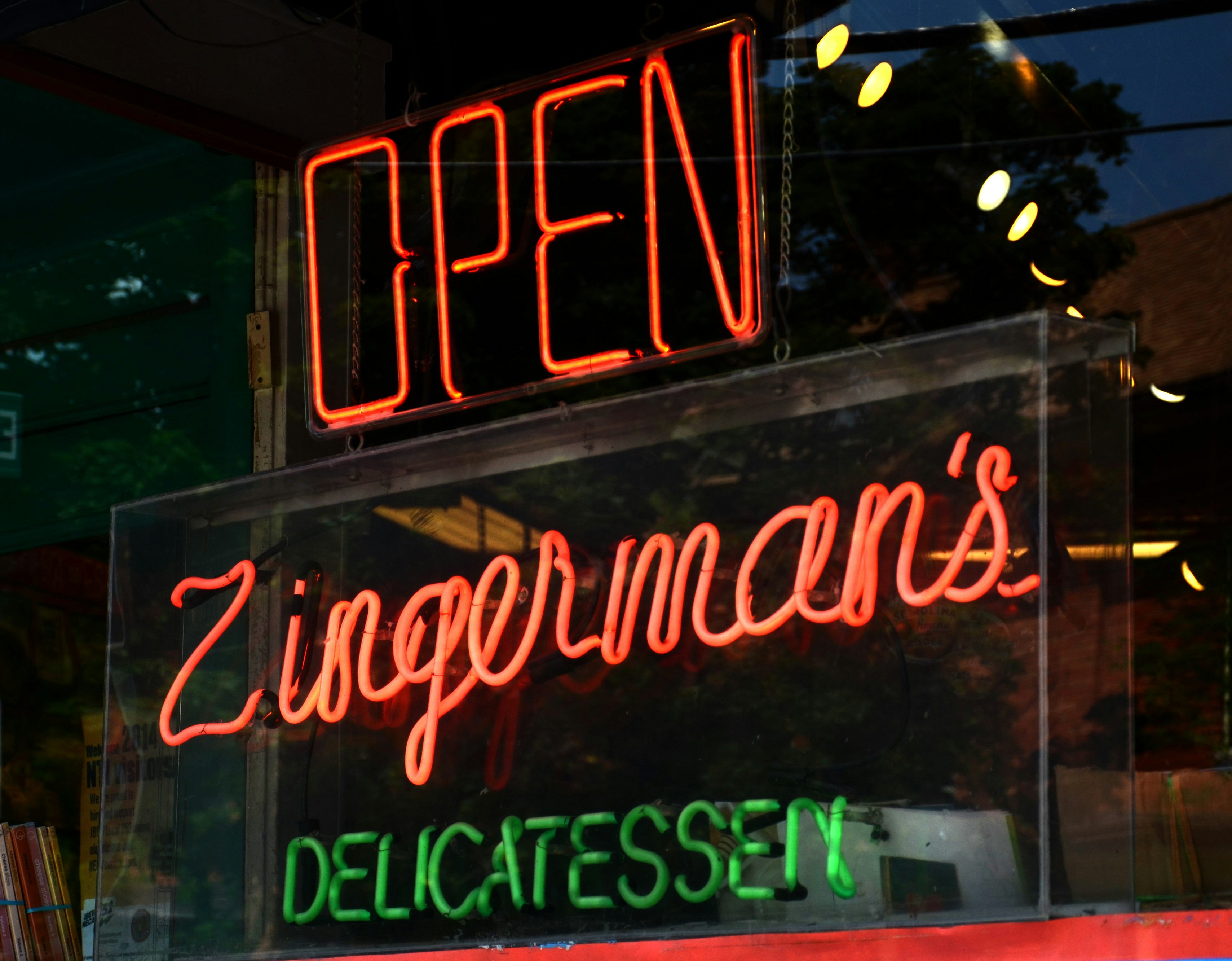 A neon sign with the words "Open, Zingerman's Delicatessen" glows in a store window; Columbus vs Ann Arbor