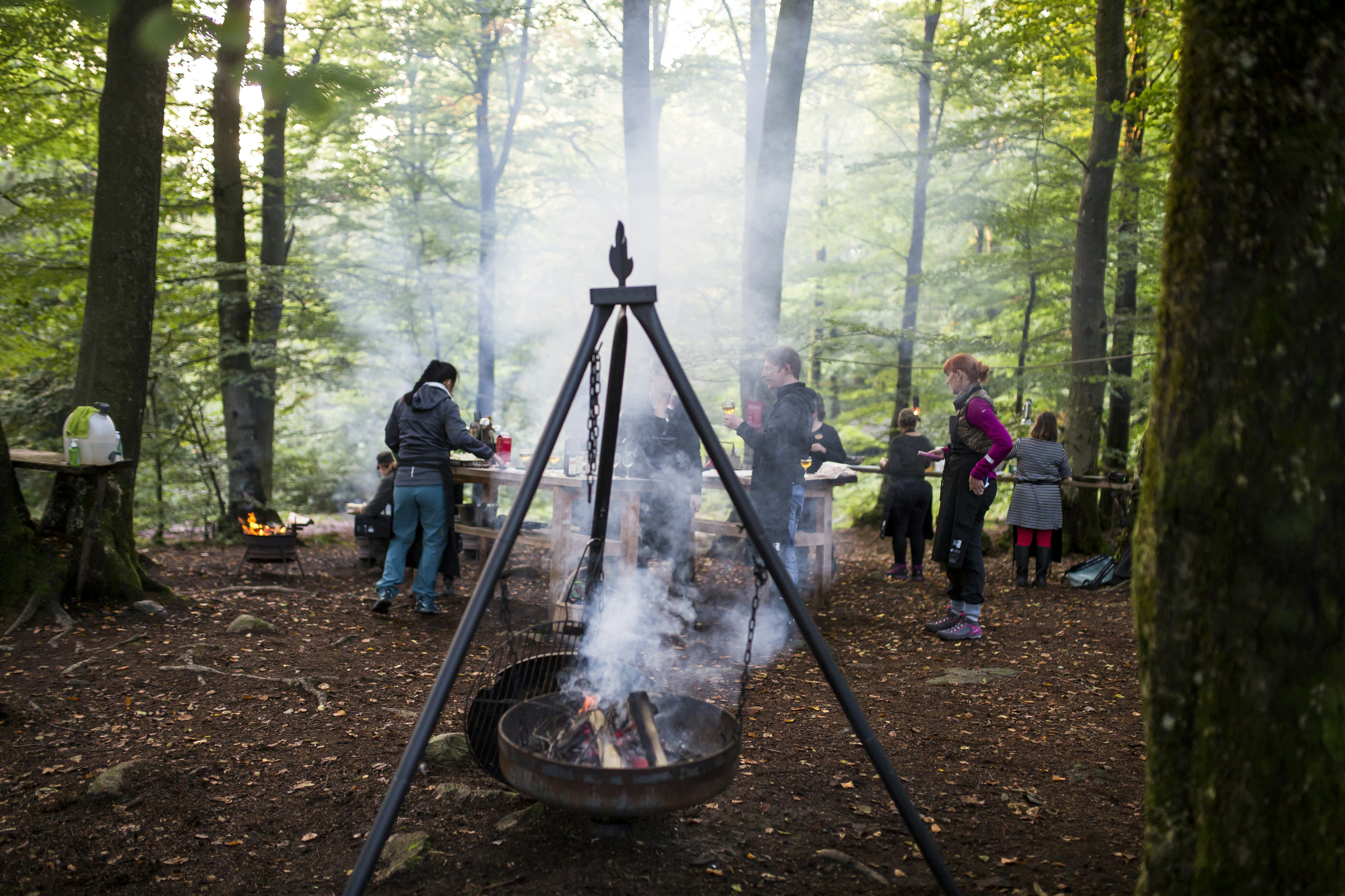 A group of eight to ten gathers around wooden tables in a forest grove, holding beverages and watching the smoke rise from a large metal cooking tripod in the center of the frame. Suspended from the tripod by three chains is a metal fire pan with burning logs. In the distance, another raised fire pit burns brightly.