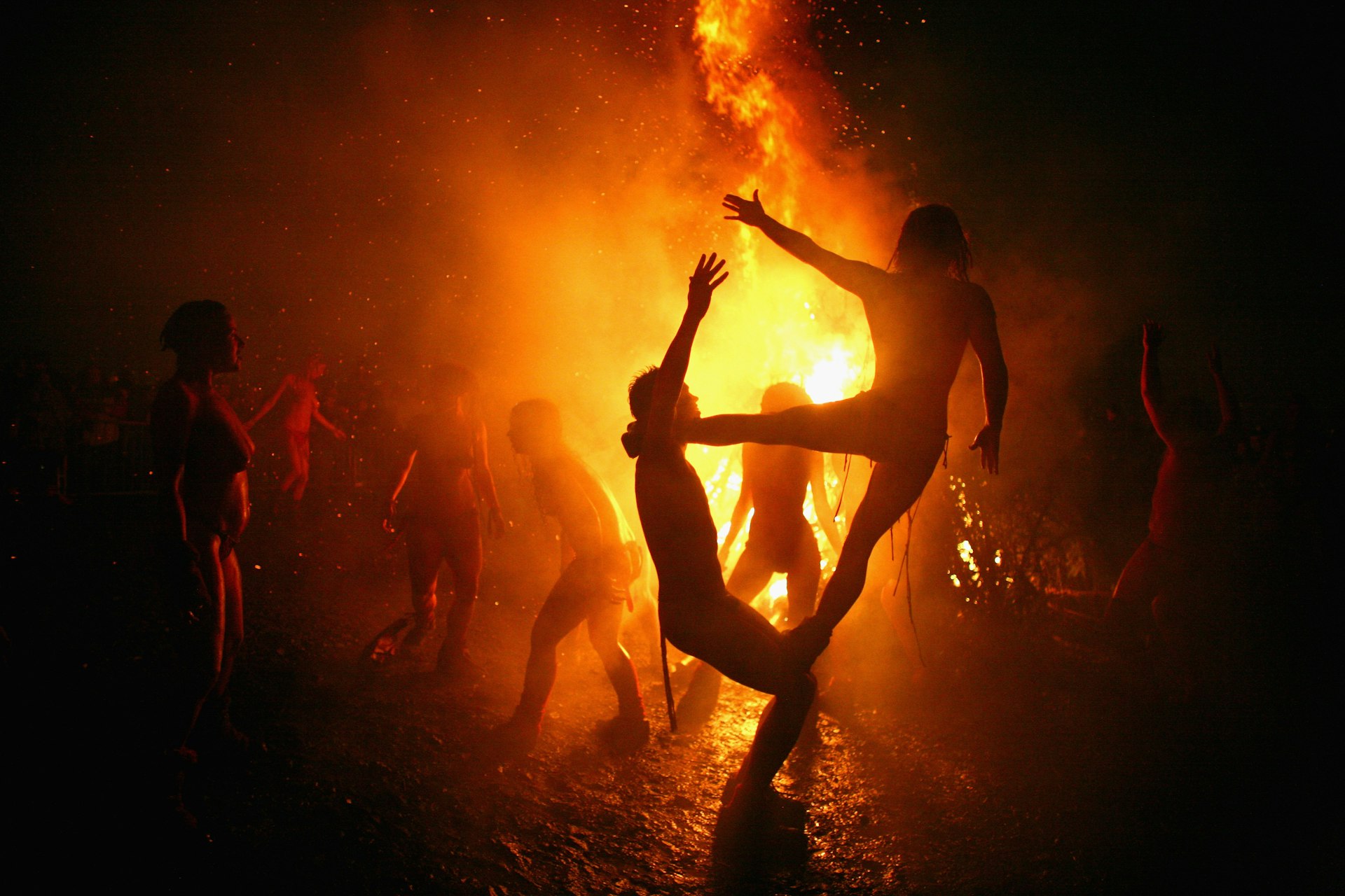 Acrobats dressed in loin cloths and body paint are in silhouette against a large bonfire. One balances their right foot on a partner's bent knees and the other leg on his shoulder, holding their arms out towards the fire as other performers dance in the background 