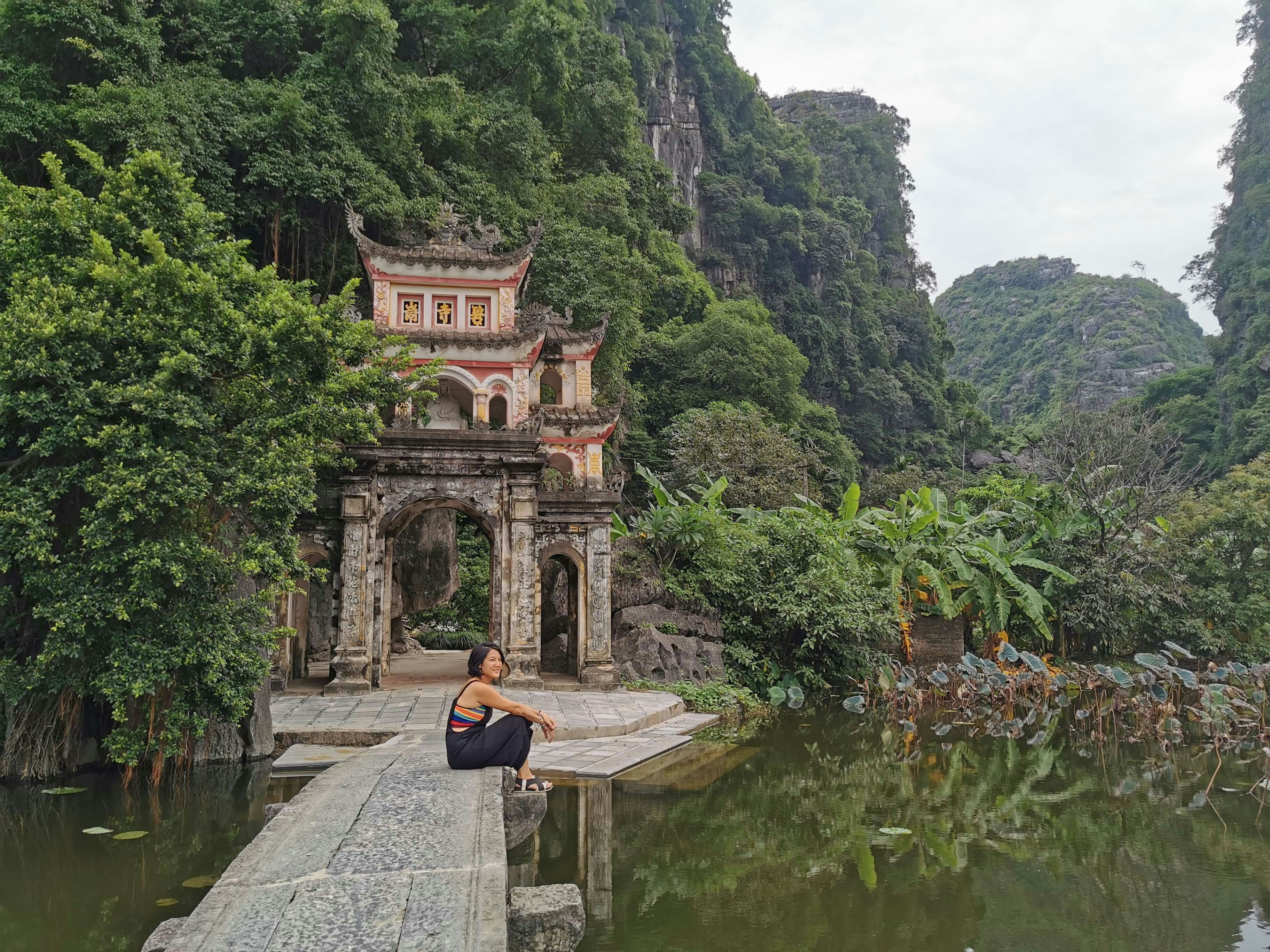 The writer, a young woman, sits on a boardwalk overlooking a river. There is a lush jungle and the remains of a temple gate in the background.