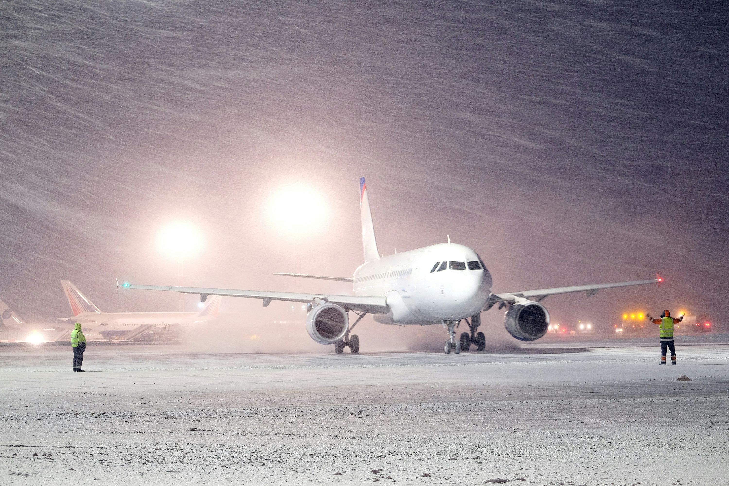 A plane sits on snow-covered tarmac at an airport at night. Two ground-staff in hi-vis vests are standing in beside it in the midst of a blizzard.