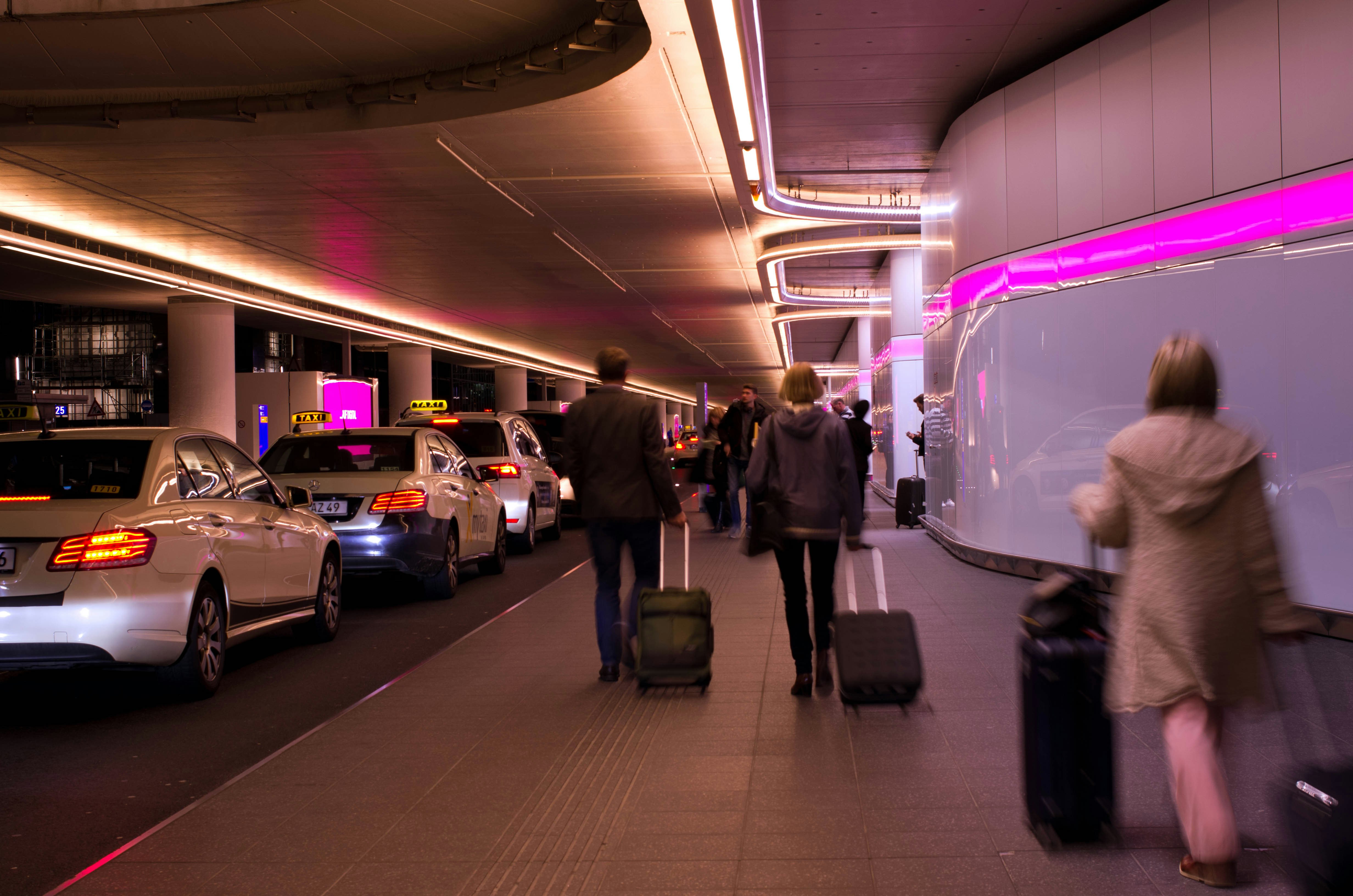 A queue of taxis lined up in an underground taxi rank at an airport; a stream of arriving passengers with suitcases are walking towards to head of the queue.